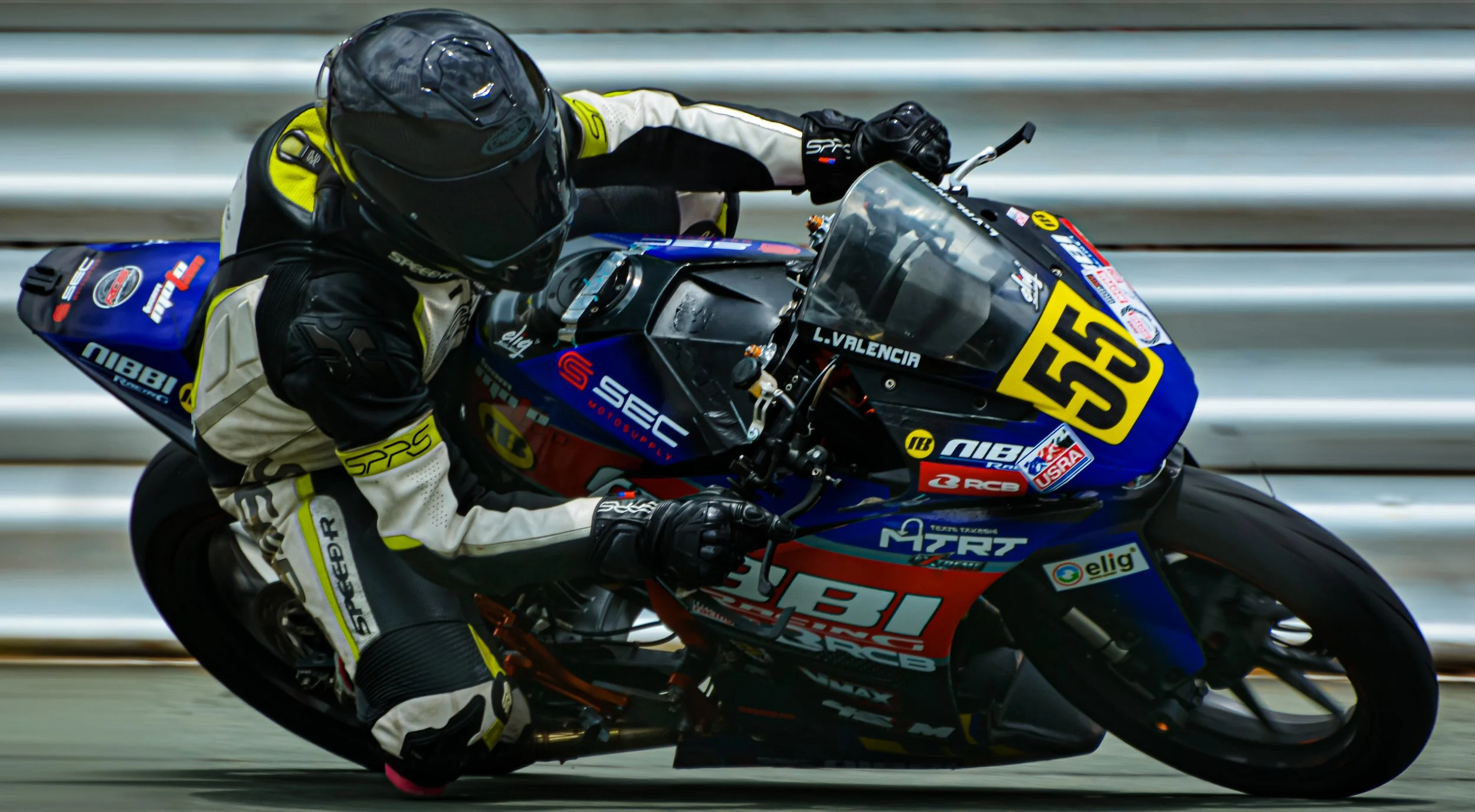 A motorcycle racer in a black helmet and racing suit leaning into a turn on a racing motorcycle with the number 55. The motorcycle has blue, black, red, and yellow colors with various sponsor logos on it, including NIBBI, AIBI, and ECS.