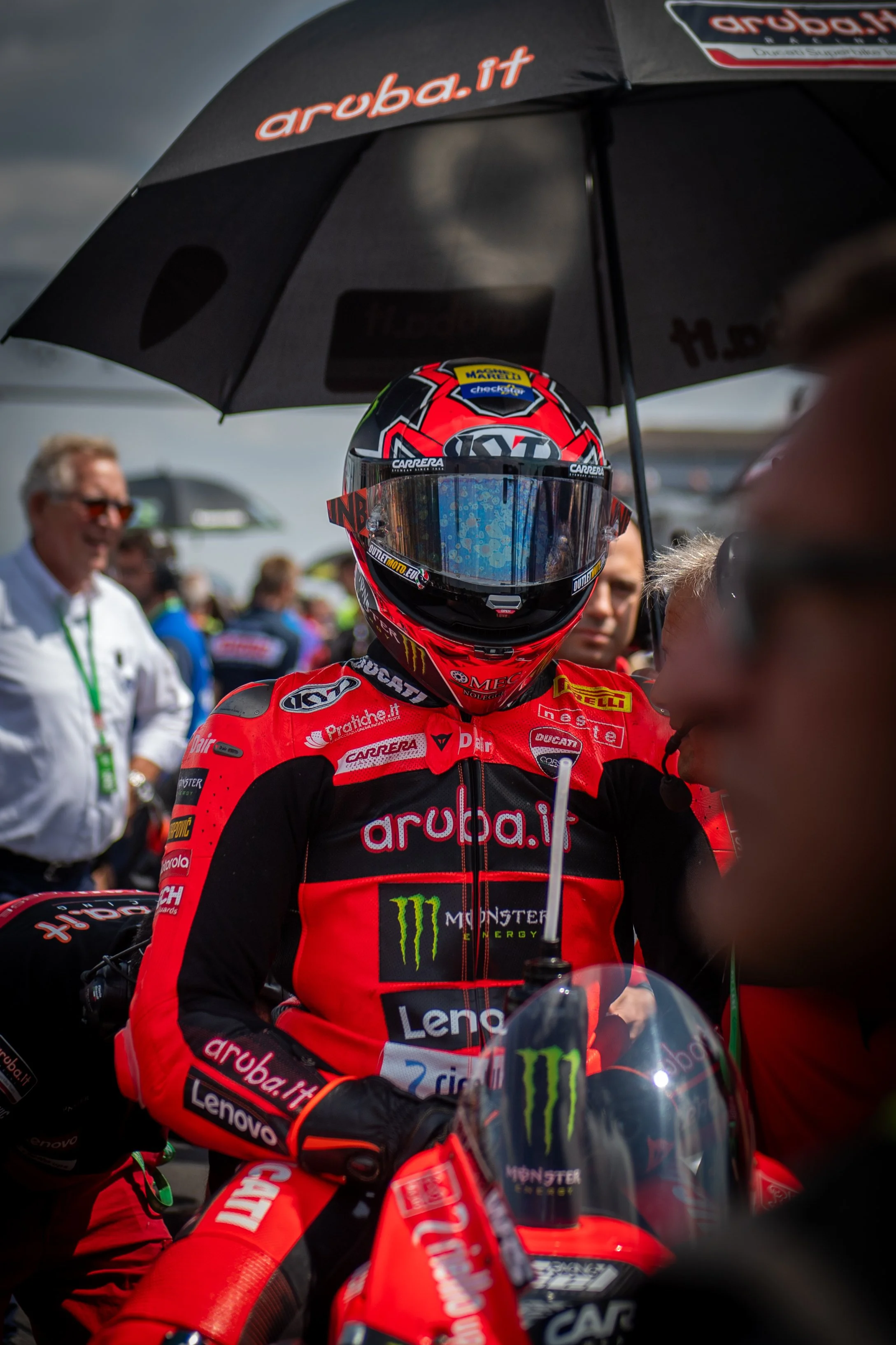 Race car driver in red racing suit and helmet standing beside Ducati motorcycle during a motorsport event, shaded under a black umbrella with 'aruba.it' branding, surrounded by team members and spectators.