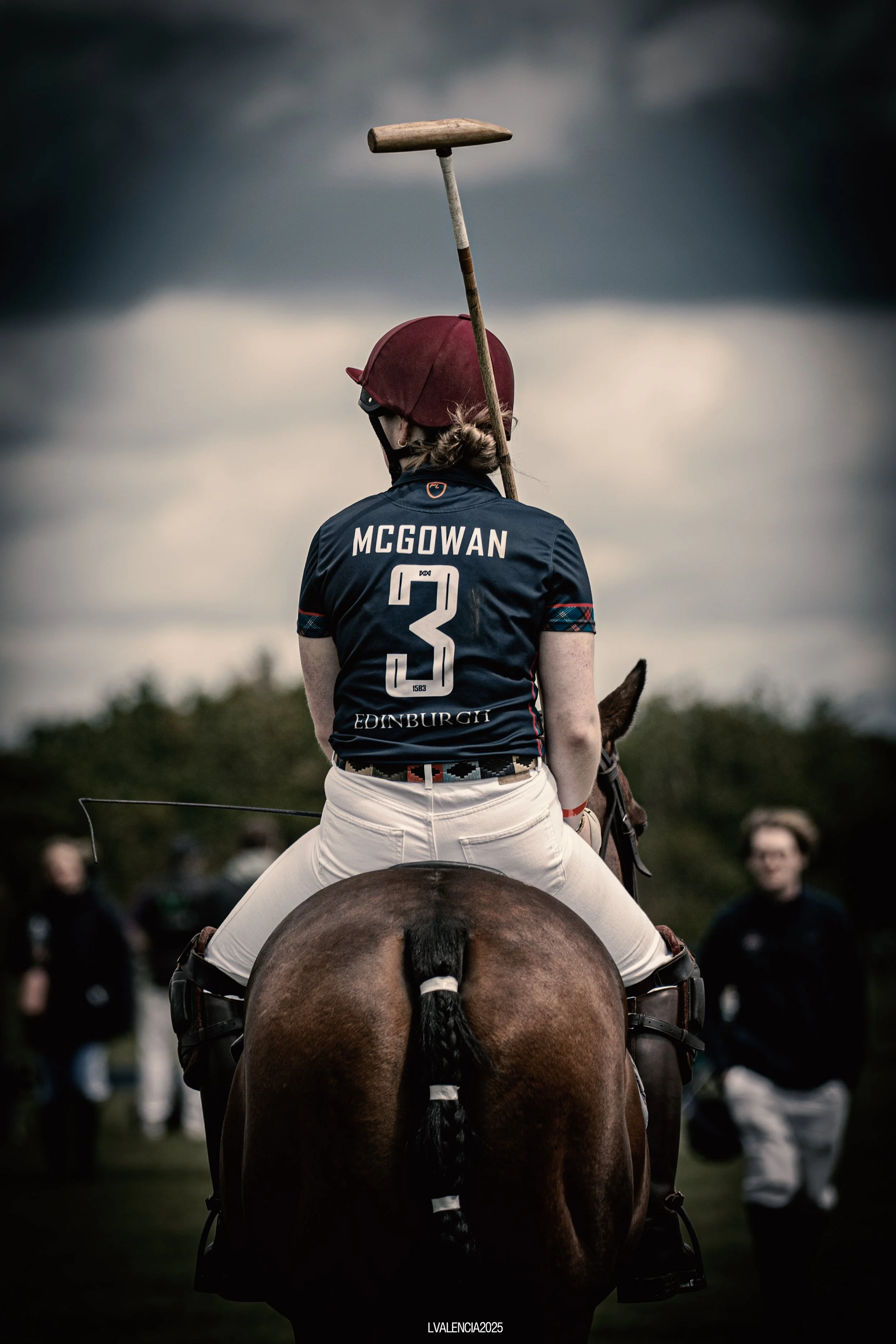 A polo player wearing a maroon helmet and a dark blue jersey with the name 'McGowan' and the number 3, sitting on a horse holding a mallet, with other people in the background under a cloudy sky.