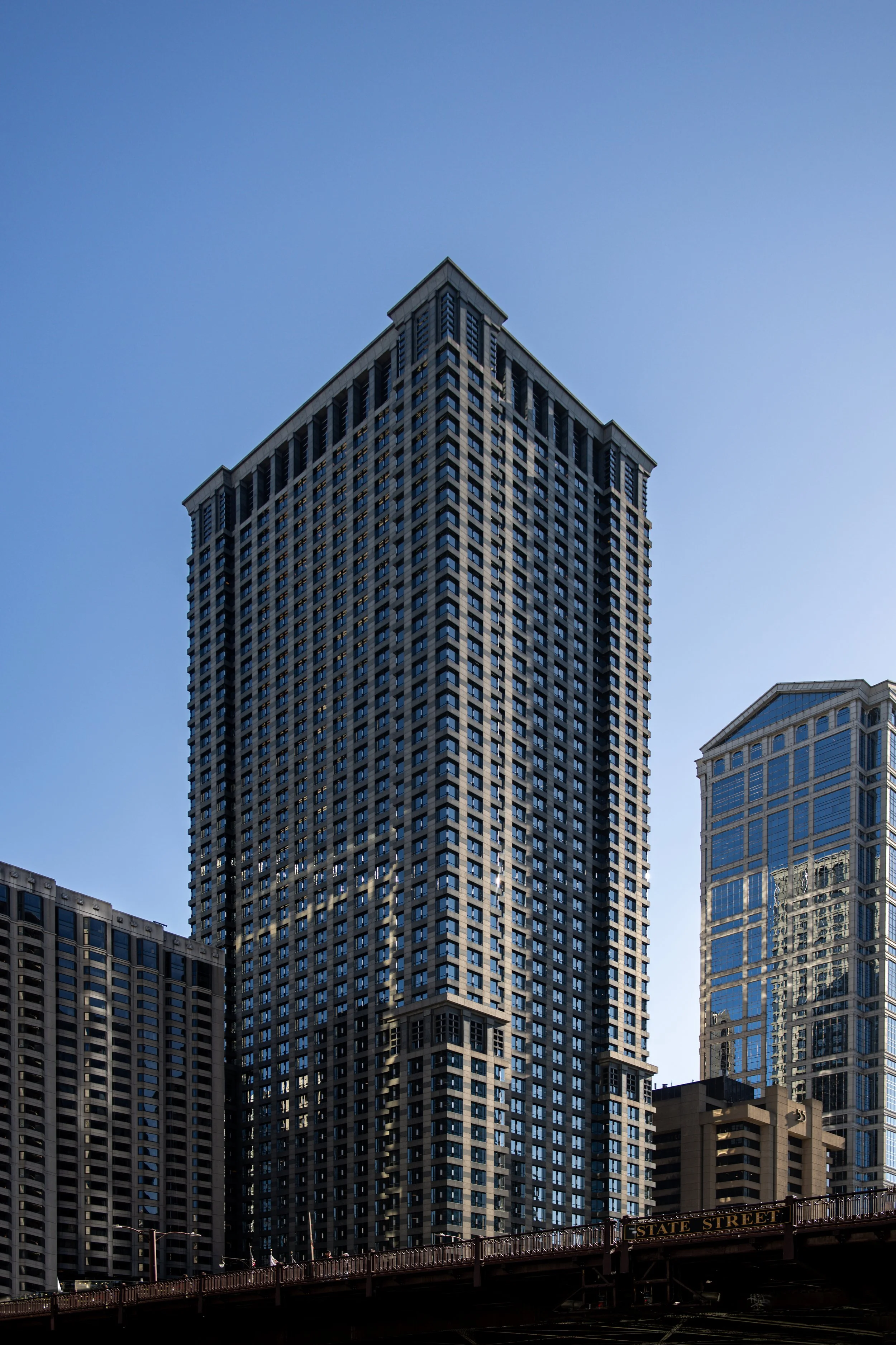 Tall modern skyscraper in an urban cityscape under a clear blue sky, with other high-rise buildings nearby and a bridge at the bottom.