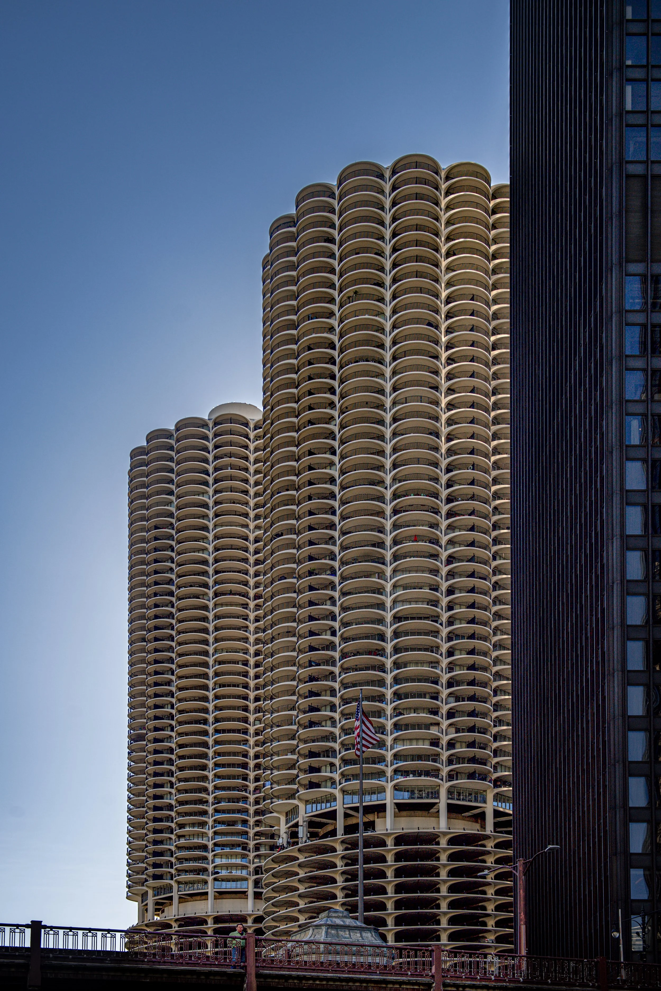 High-rise building with rounded balconies stacked in a column, adjacent to a dark tall building, with a flagpole and American flag at the base, under a clear blue sky.