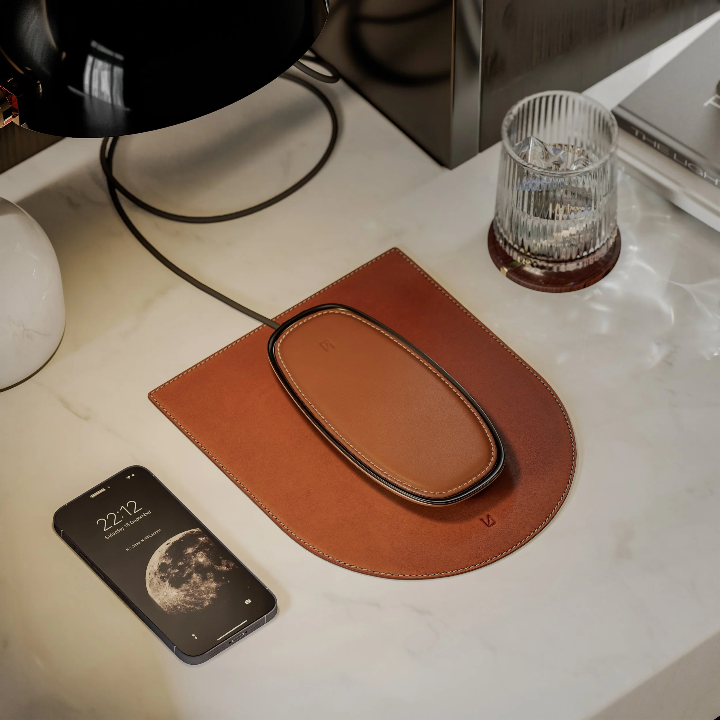 A desk with a smartphone displaying a moon background, Lunar Artefacts wireless charger on a leather desk mat, a glass of water, a book, and a desk lamp.