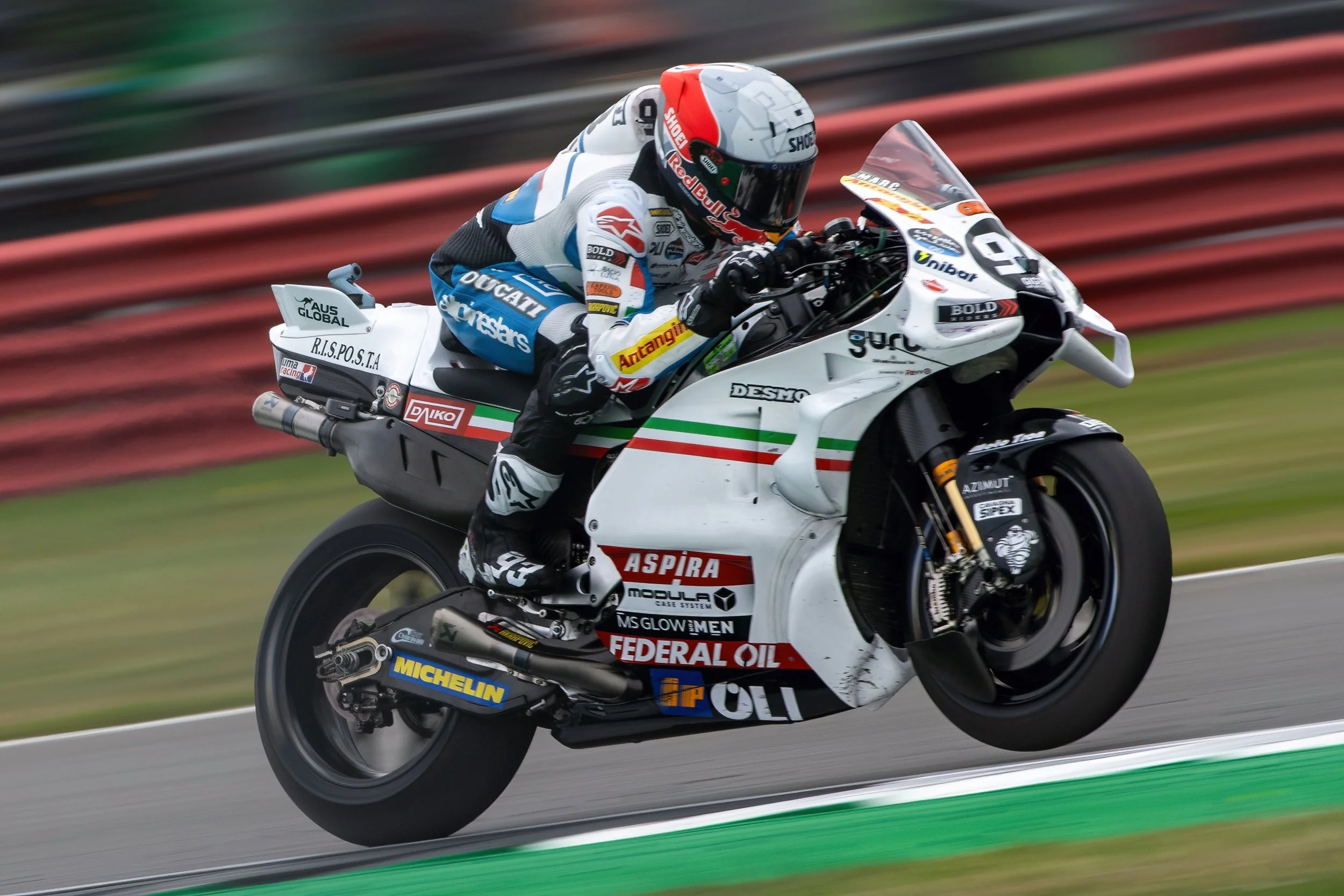 A motorcycle racer in racing gear riding a white sportbike on a race track, with a blurred background of red and green barriers.