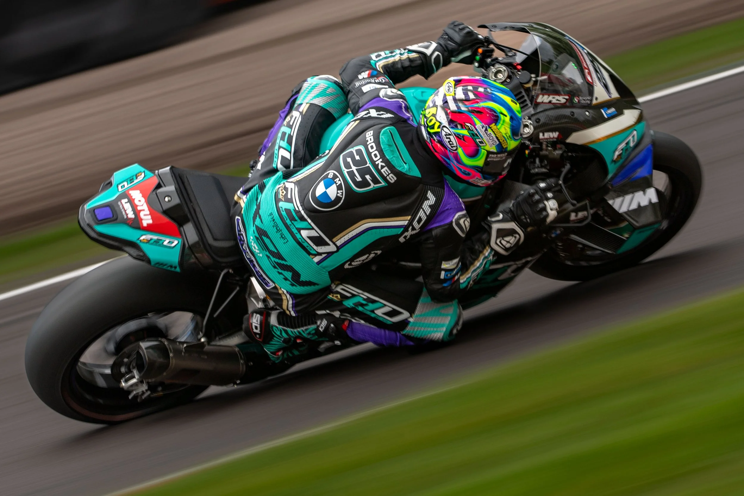 Motorcycle racer leaning into a turn on a racetrack, wearing a racing suit and a colorful helmet, with a blurred background.