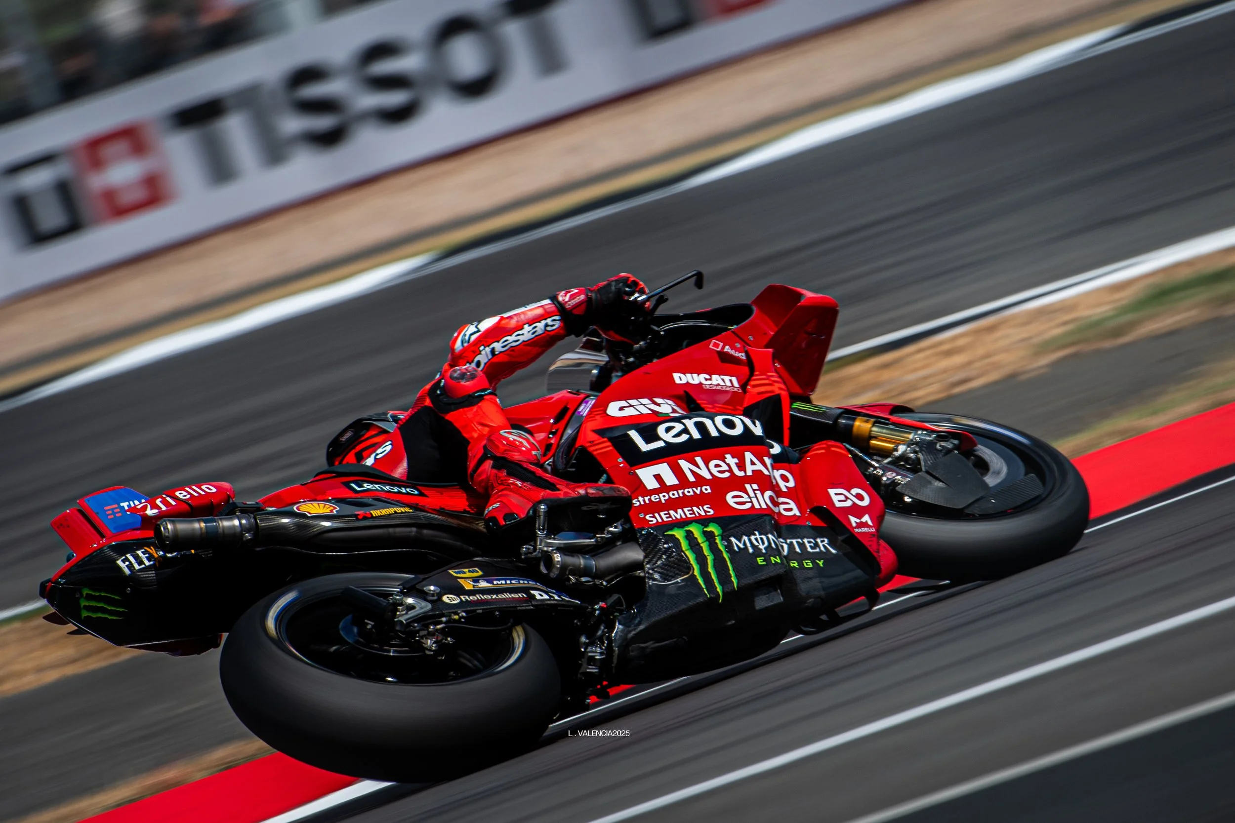 A professional motorcycle racer wearing red gear is leaning into a turn on a red and black Ducati race bike on a racetrack.