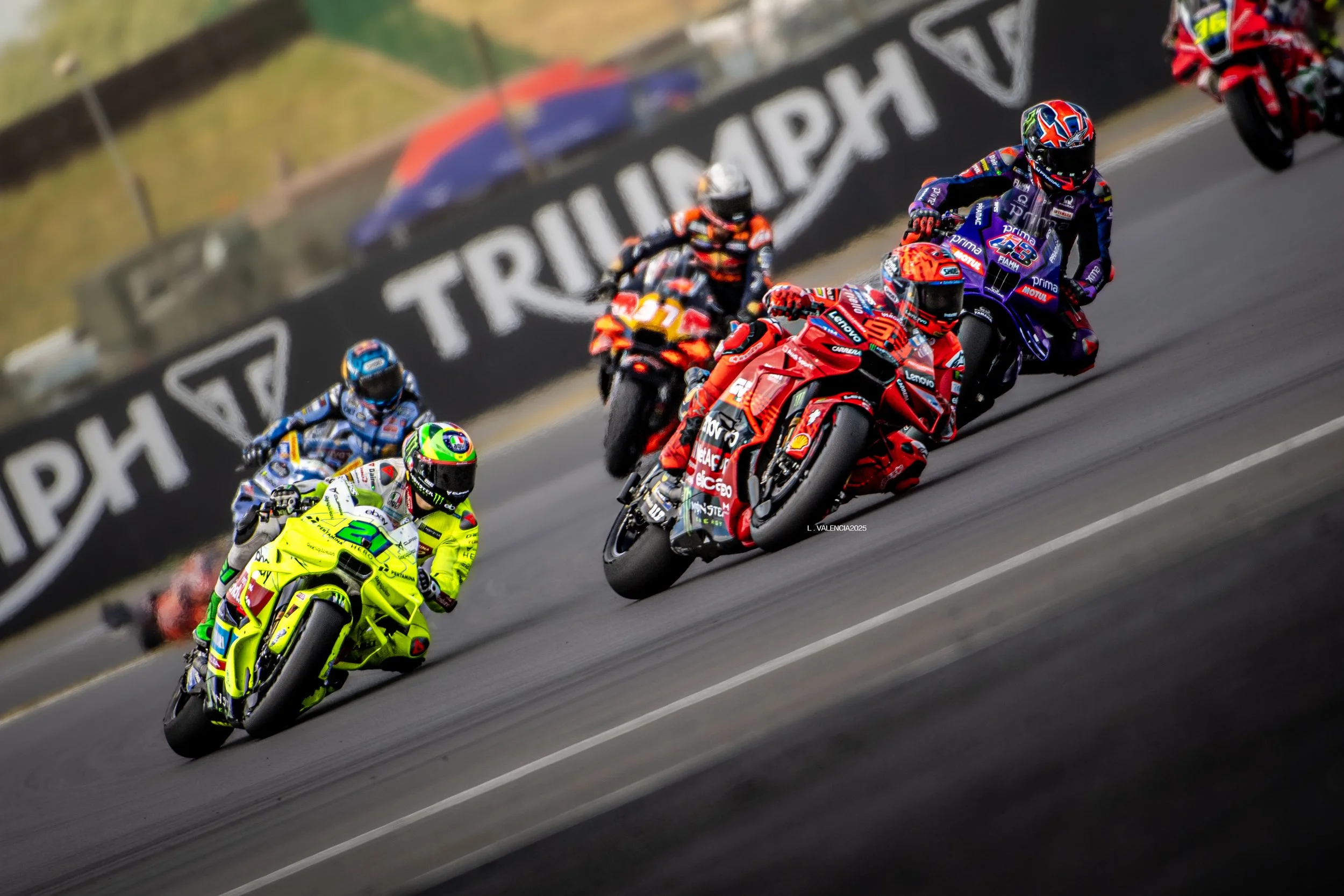 Motorcycle racers wearing colorful gear leaning into a turn on a racetrack, with a banner in the background.