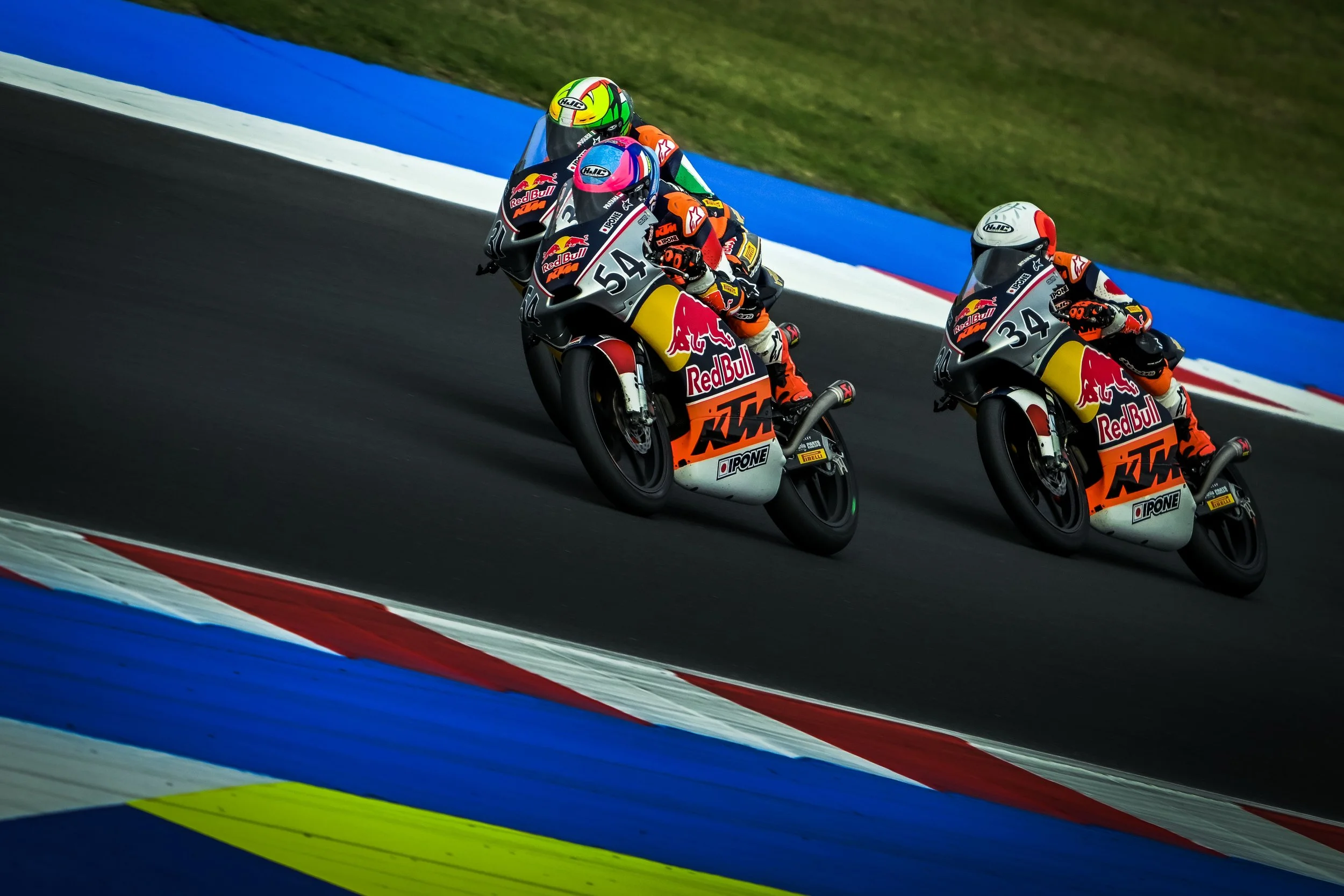 Two motorcycle racers in colorful gear riding on a racetrack with blue, red, white, and yellow painted sections.