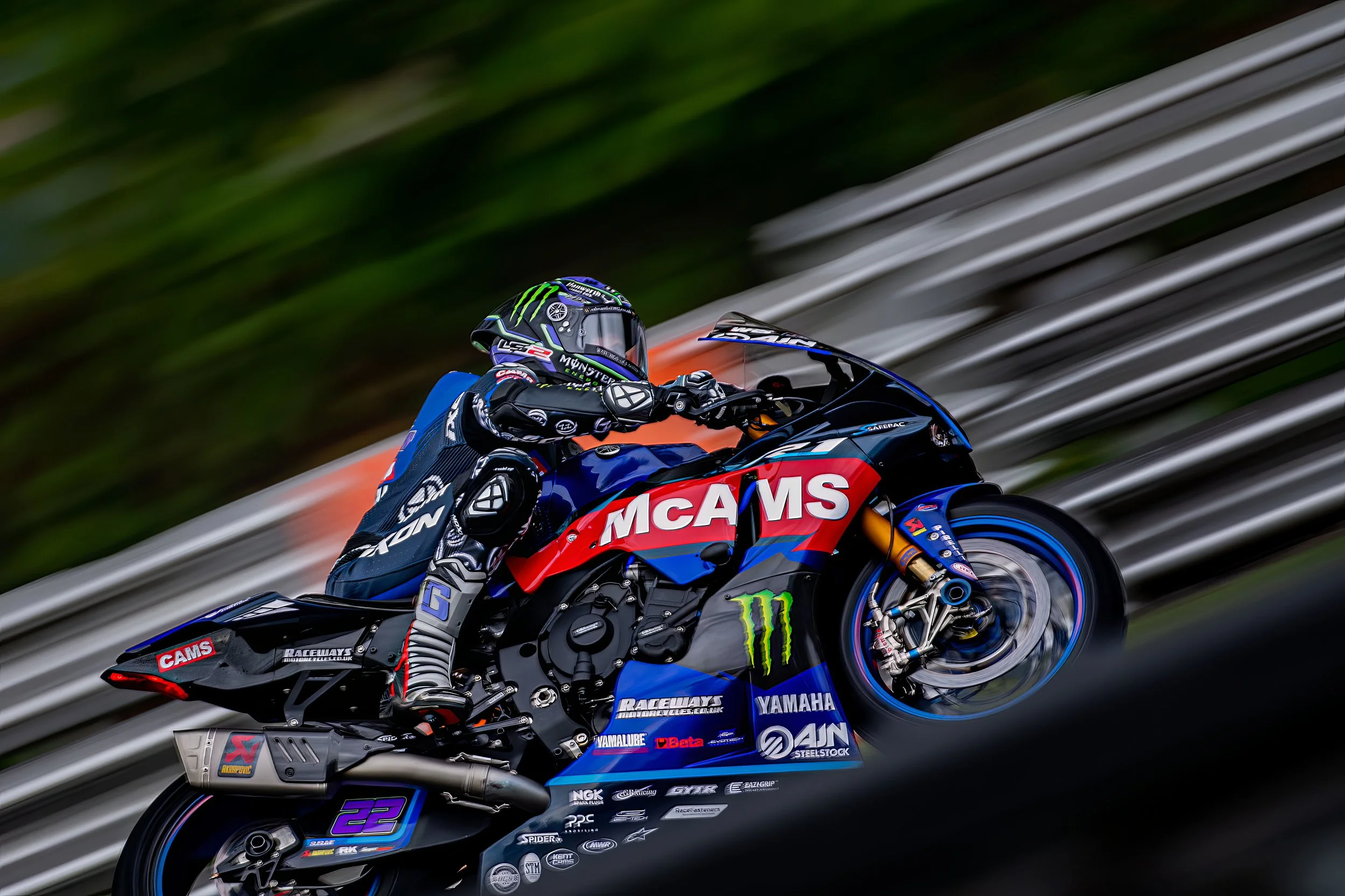 Motorcycle racer in full gear riding on track with a black, red, and blue motorcycle branded with McAMS, Monster Energy, Yamaha, and other logos, blurred background of greenery and track barriers.