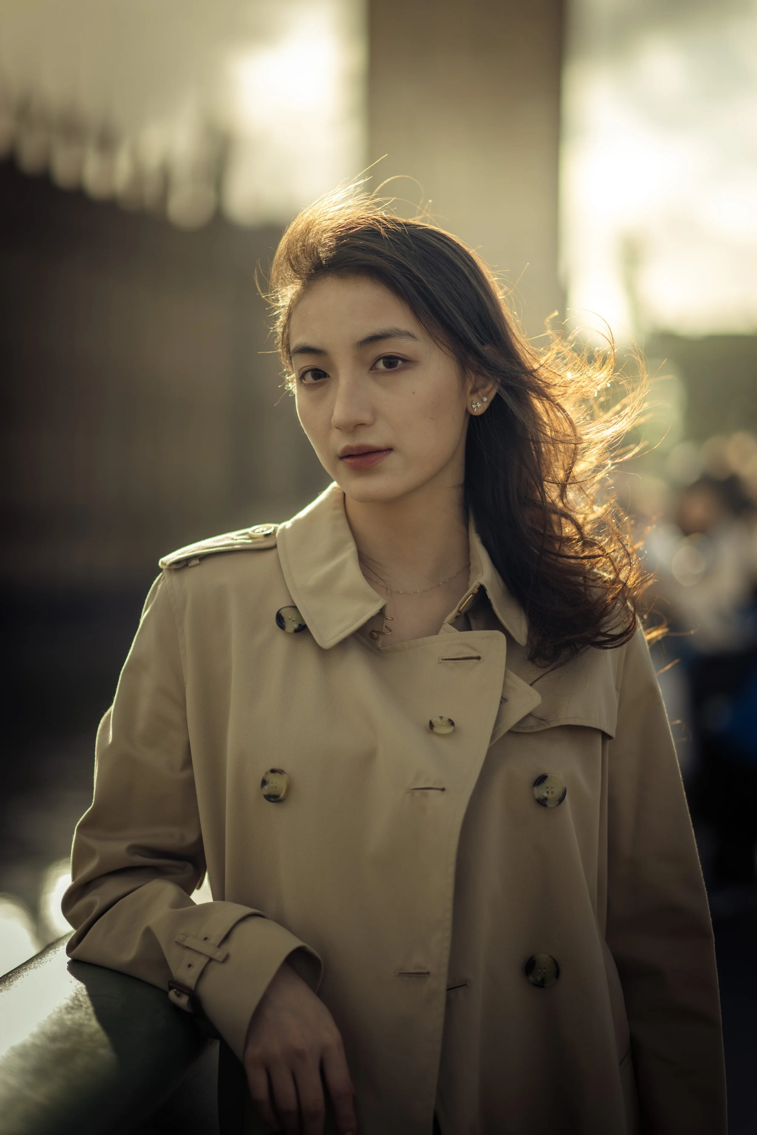 A young woman wearing a beige trench coat, standing outdoors with sunlight behind her and city scenery in the background.