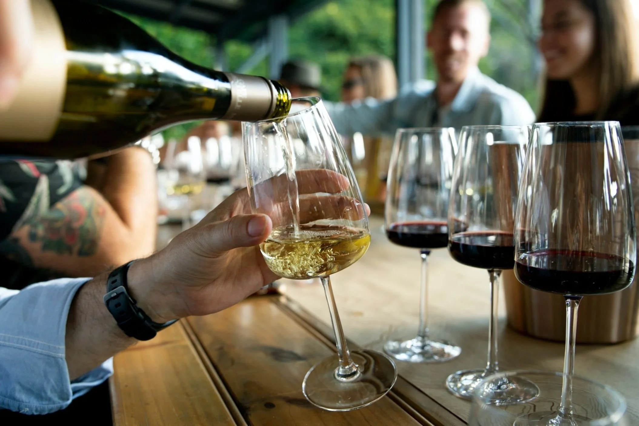 A person pouring white wine into a glass at a table with several glasses of red wine and people socializing in the background.