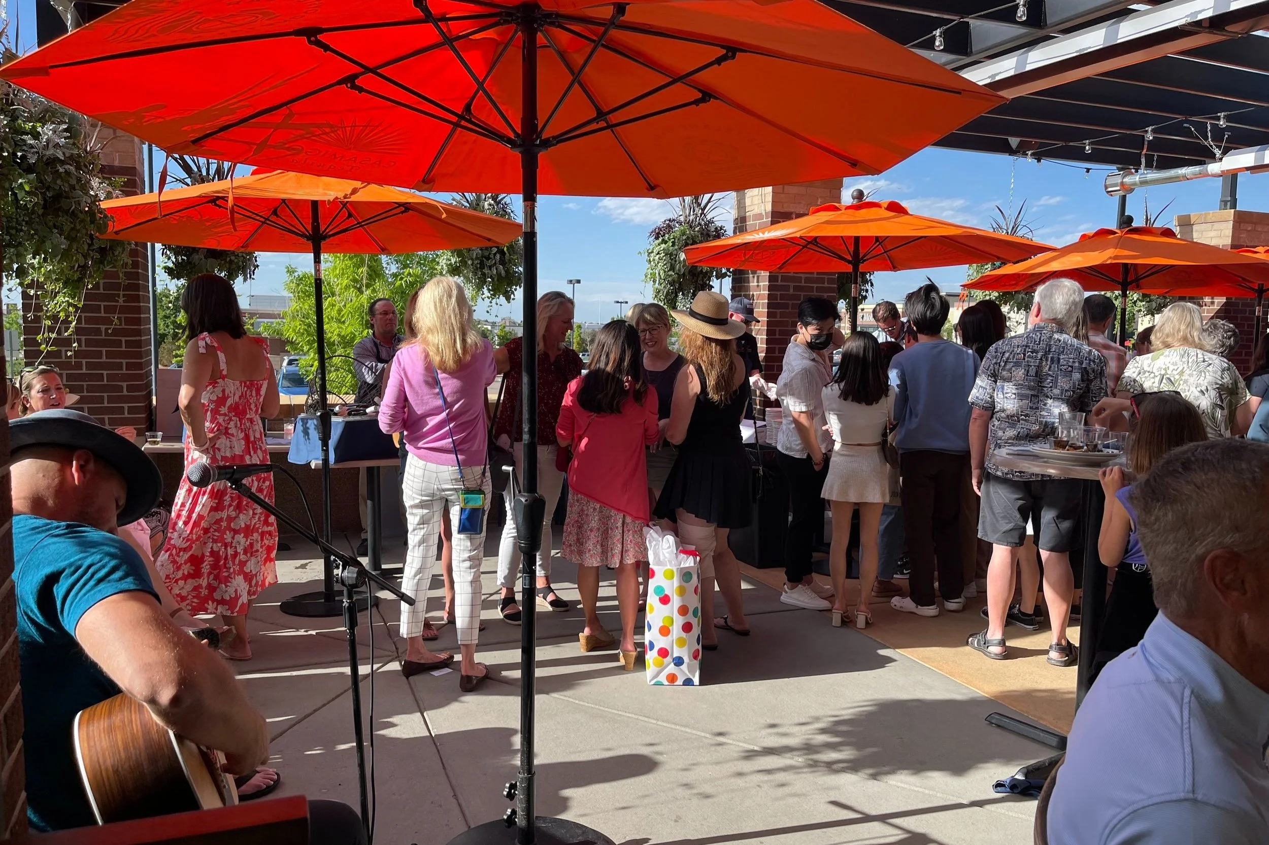 A lively outdoor gathering at a restaurant patio with several people socializing and mingling under bright orange umbrellas. A musician is playing an acoustic guitar near a microphone in the foreground. People are dressed in casual, summery attire and the scene is sunny with green plants in the background.