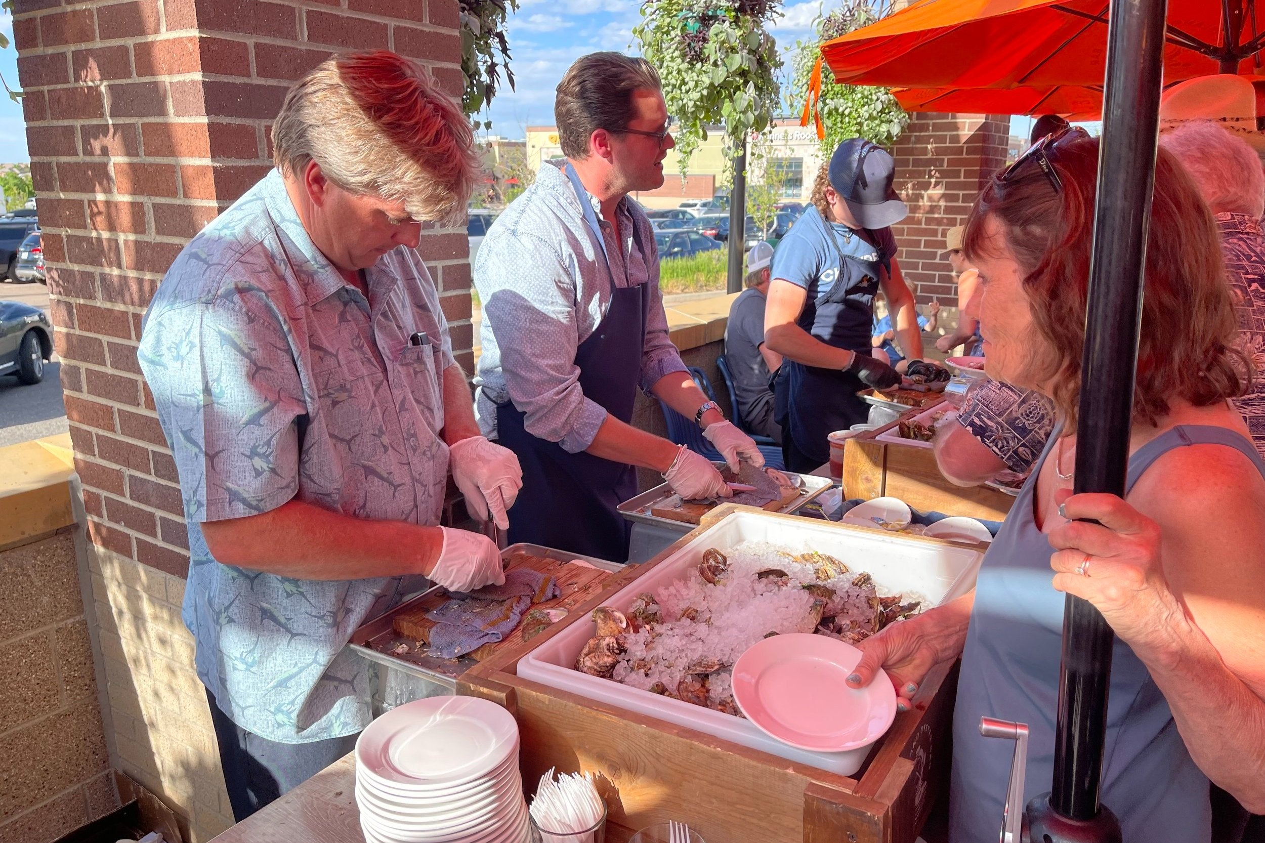 People shucking oysters at an outdoor seafood event near a brick wall with plates and ice.