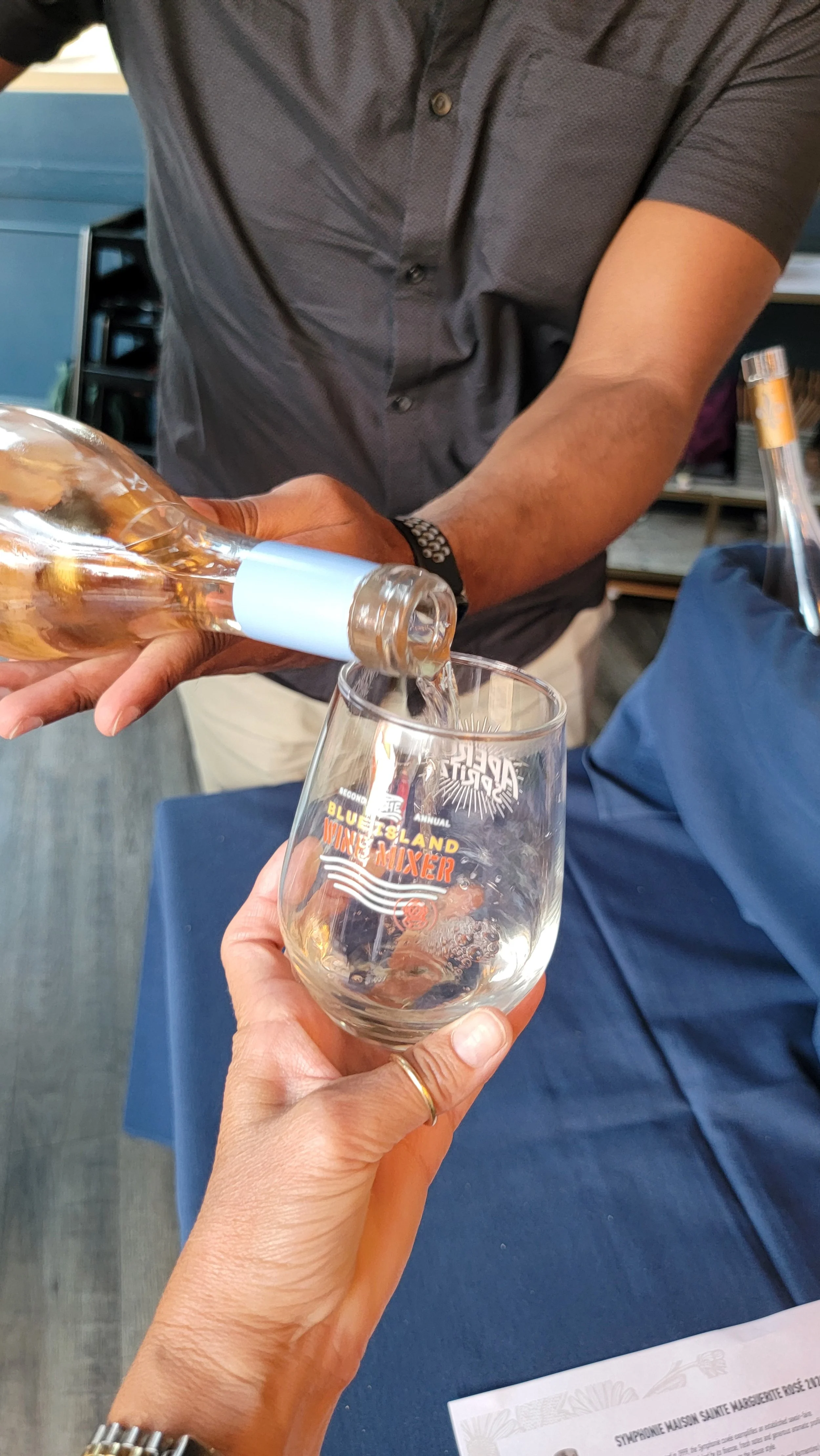 A person pours a drink from a clear bottle into a glass with a logo that reads 'Bluesland Wine Mixer' on a table, with another person holding the glass. The person pouring is wearing a dark gray button-up shirt, and the person holding the glass has a gold ring on their finger.