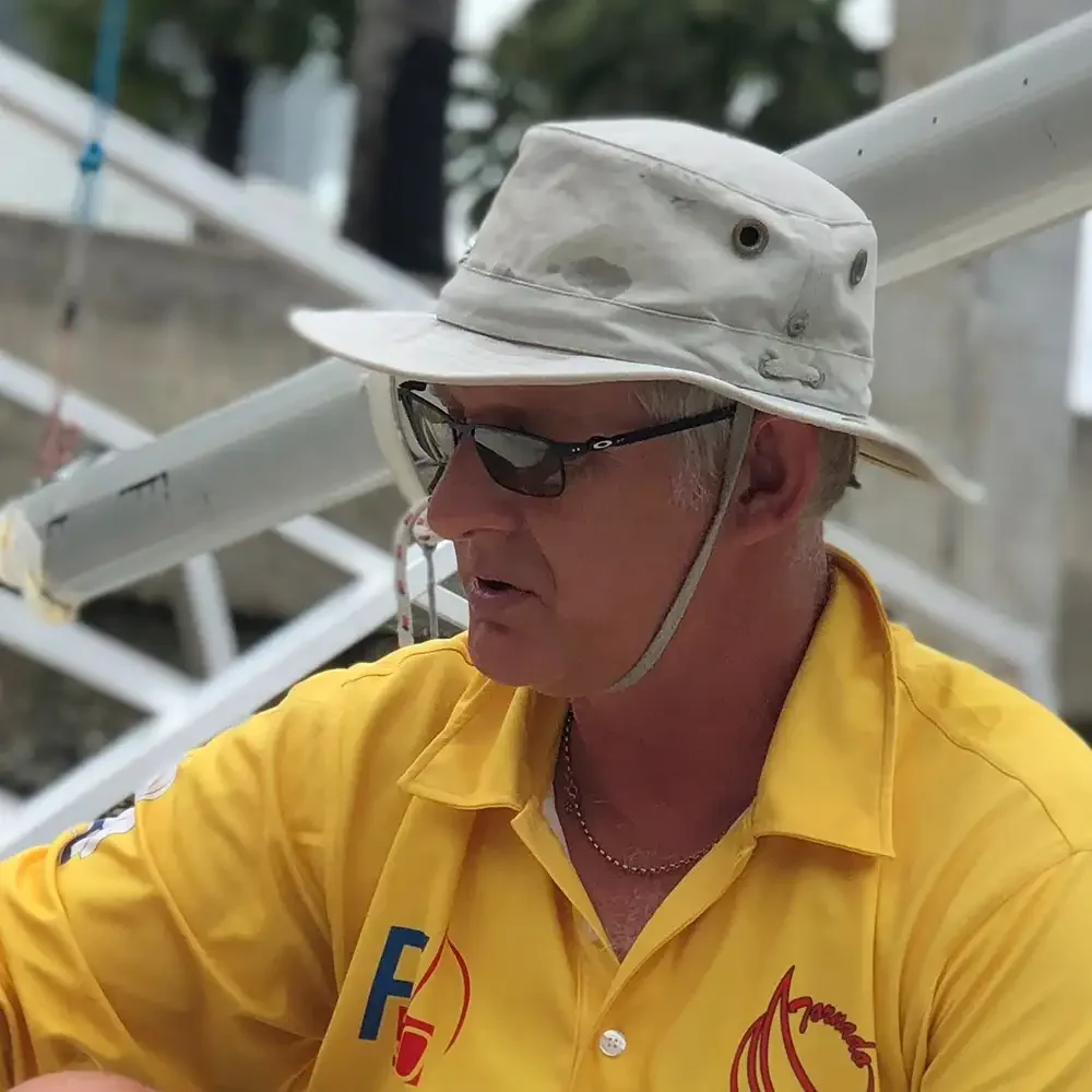 Portrait headshot photo of Nic Wymer, an older man wearing a yellow shirt, sunglasses, and a wide-brimmed hat with vents, outdoors, near white metal structures.