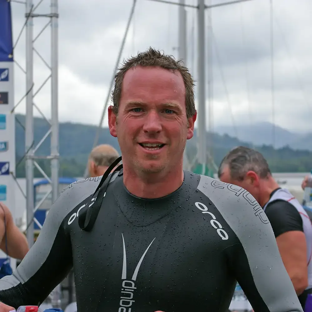 Portrait headshot photo of Ed Curtis, smiling man in a wet black and gray Triathlon wetsuit, with other people and a cloudy sky in the background.