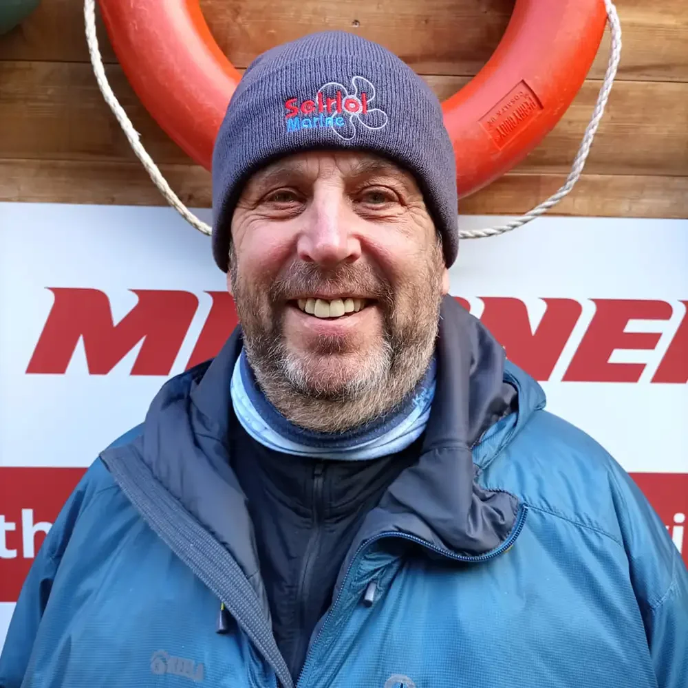 Portrait headshot photo of Neil Rawlinson, a middle-aged man wearing a gray beanie with a red and blue logo, a blue jacket, and a neck gaiter, standing in front of a life preserver.