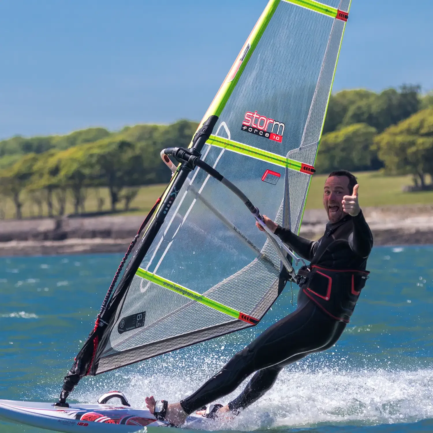 A man windsurfing on the water, giving a thumbs-up and smiling, with trees and a shoreline in the background.