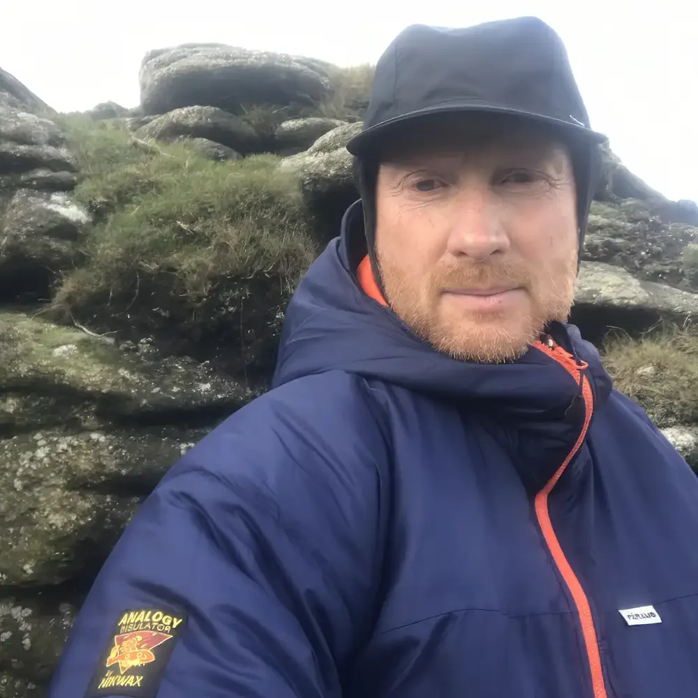 Portrait headshot photo of Kevin Found, man wearing a blue thermal jacket, and black cap, standing in front of a mossy stone wall.