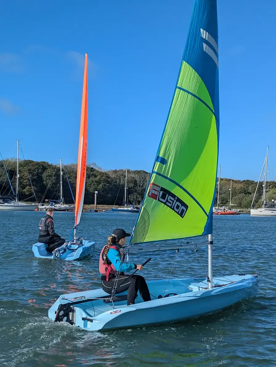 Two people sailing small sailboats on a calm body of water with other boats docked in the background and a clear blue sky above.