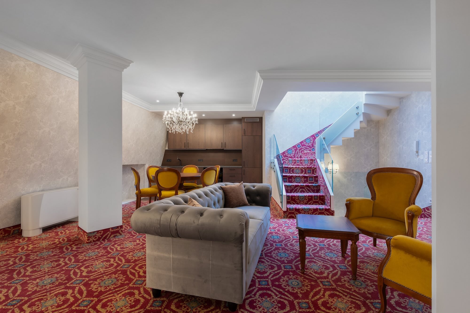 Living room with beige patterned walls, a chandelier, a grey tufted sofa, a yellow armchair, a small wooden table, a staircase with pink patterned carpet, a dining area with yellow cushioned chairs, and a wooden cabinet in the background.