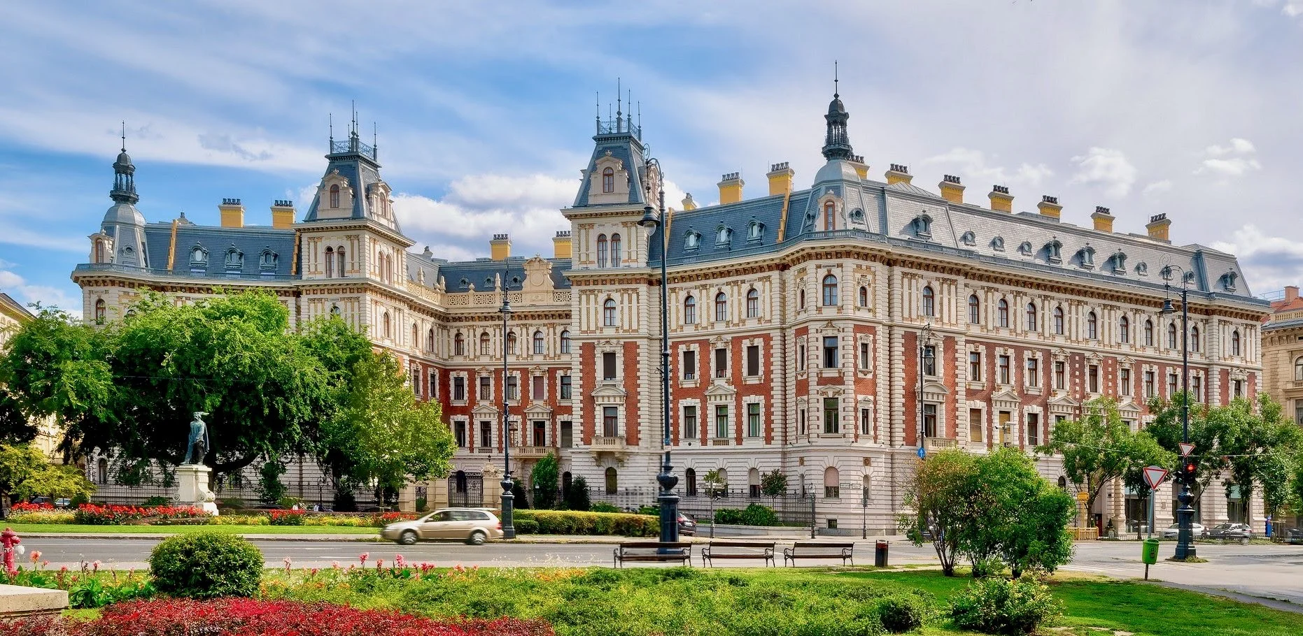 A large historic building with ornate architecture and multiple towers, seen from the street with cars passing by and a park with green trees and flowerbeds in the foreground.