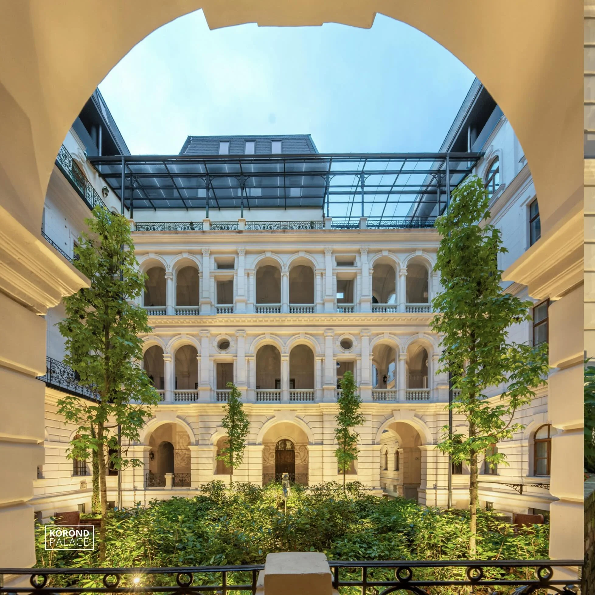 View of a classical courtyard with white five-story building surrounding it, featuring arches, balustrades, and columned balconies, green trees in the courtyard, and a blue sky above.