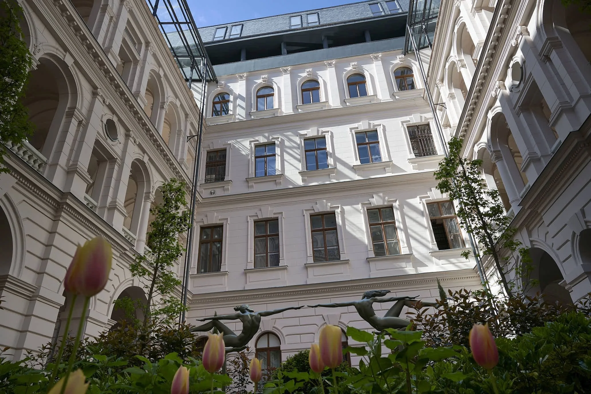 View of a courtyard with classical white architecture and rounded windows, framed by trees and flowering plants, with tall modern building visible above.