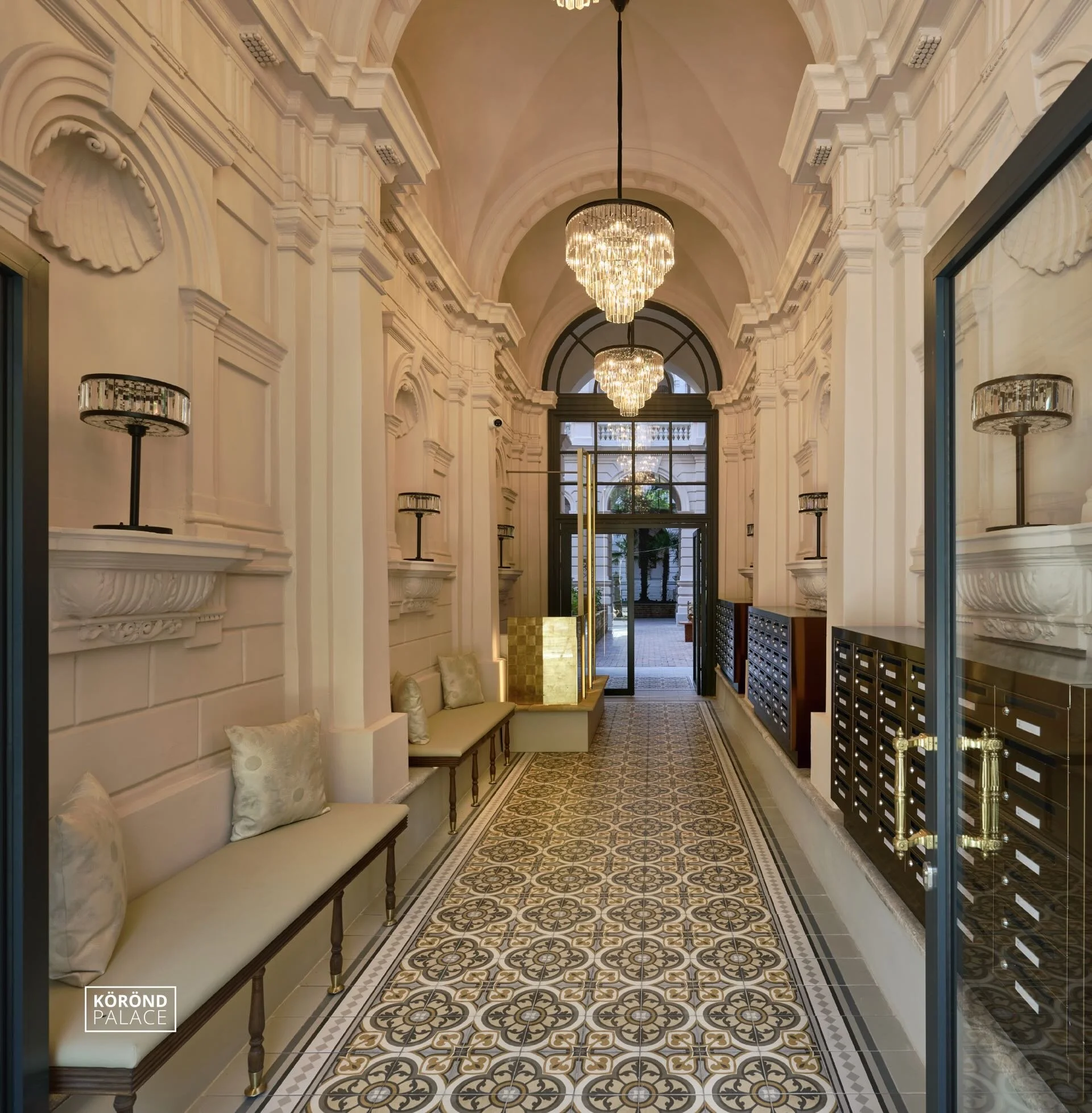Elegant hotel lobby with ornate white walls, chandeliers, and patterned tile floor, featuring seating with pillows and mailboxes lined up on the right side.