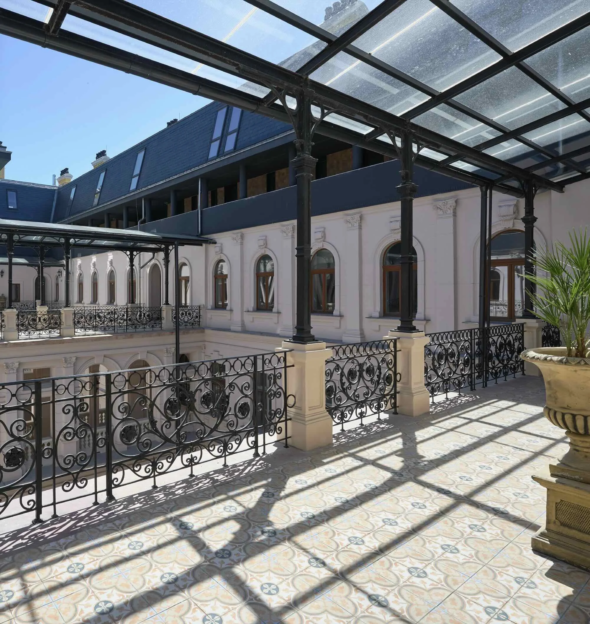 View of a spacious balcony with ornate black wrought iron railings and decorative tiles on the floor, overlooking an inner courtyard of a historic building with white walls, arched windows, and a glass canopy overhead, casting shadows on the floor.
