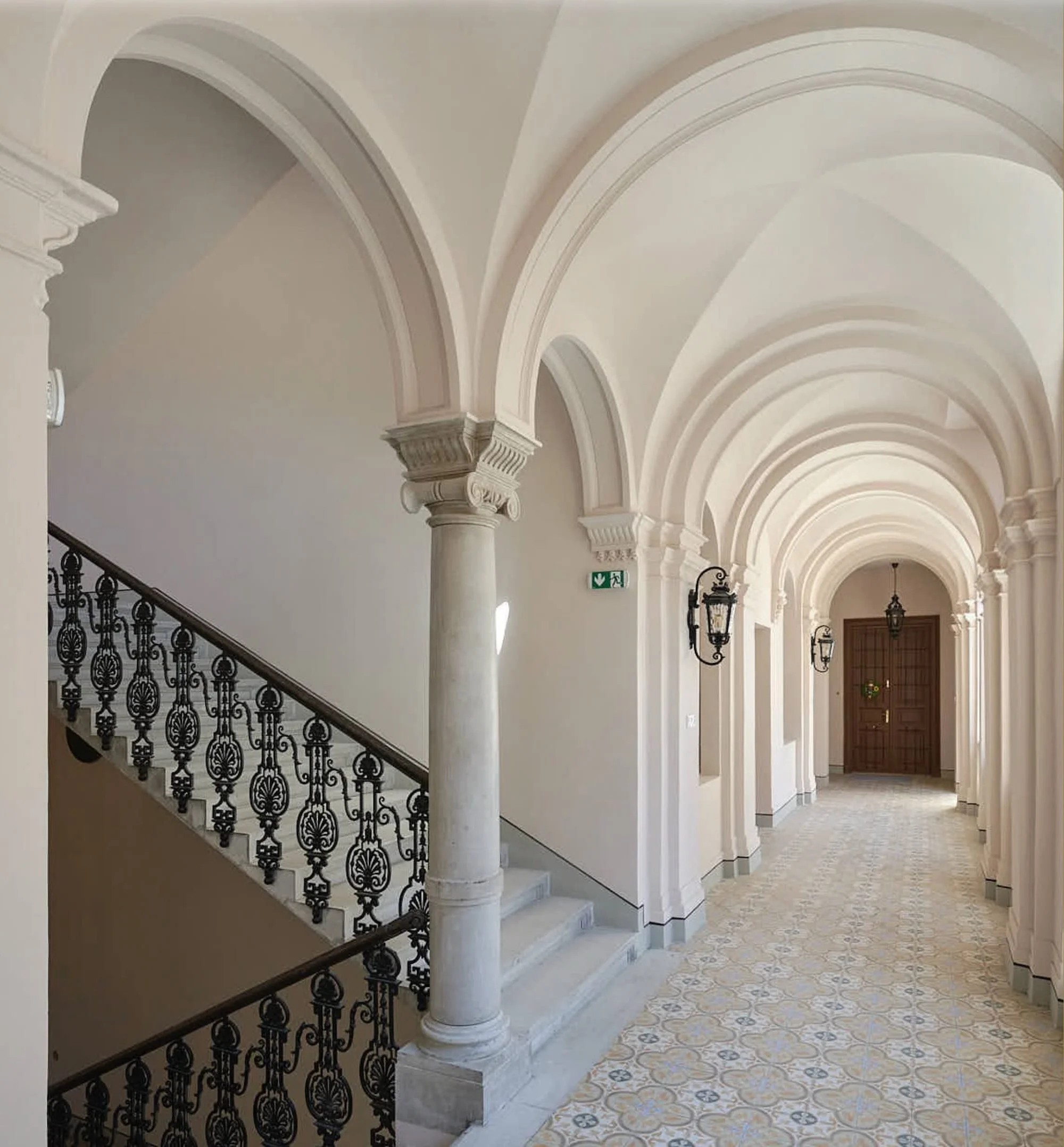 Elegant hallway with vaulted arches, white columns, ornate light fixtures, patterned tile floor, and a closed wooden door at the end.