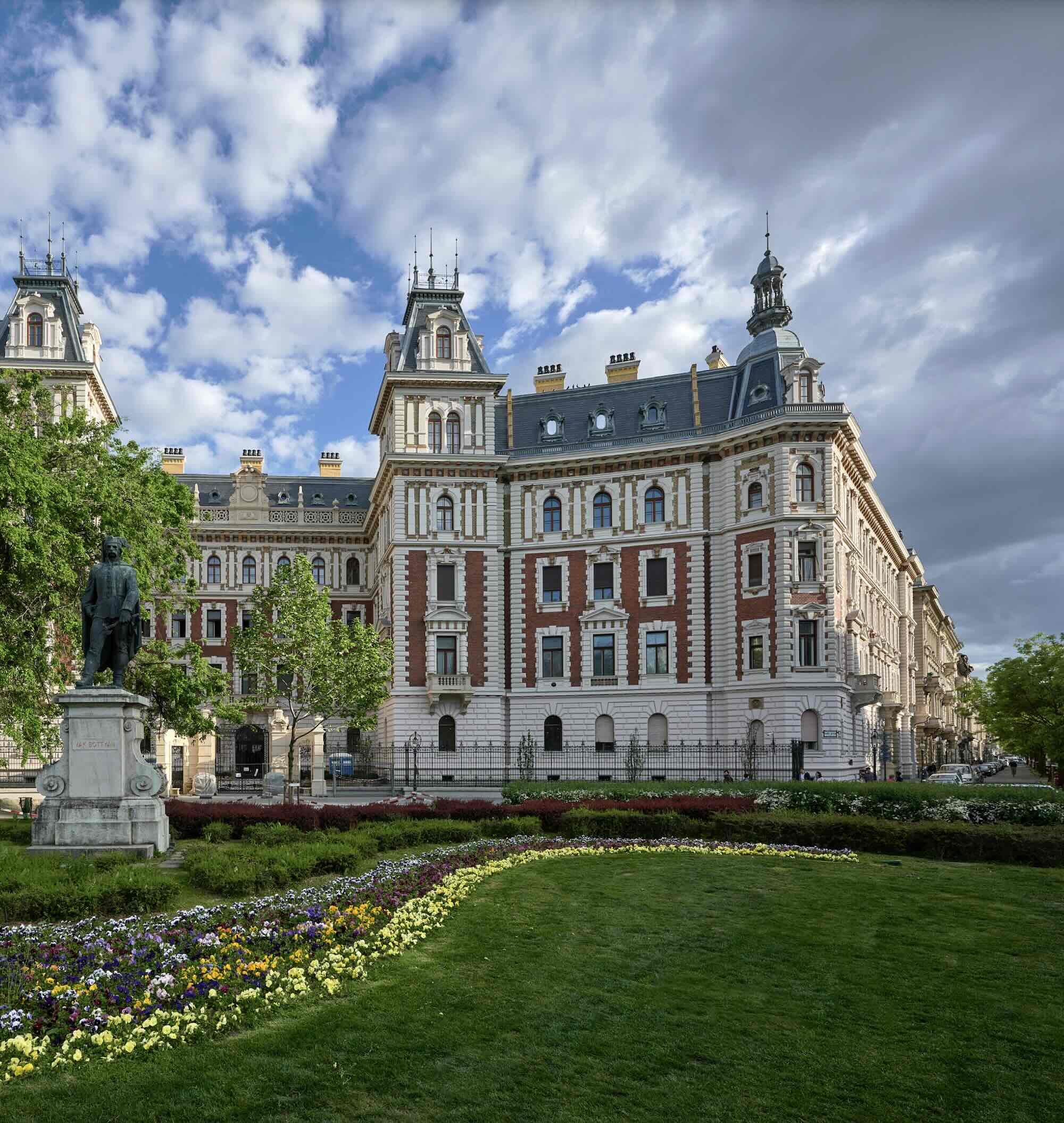 A large, historic, ornate building with towers, windows, and intricate architectural details, set against a partly cloudy sky surrounded by greenery and flowering beds, including a statue of a person on a pedestal in the foreground.