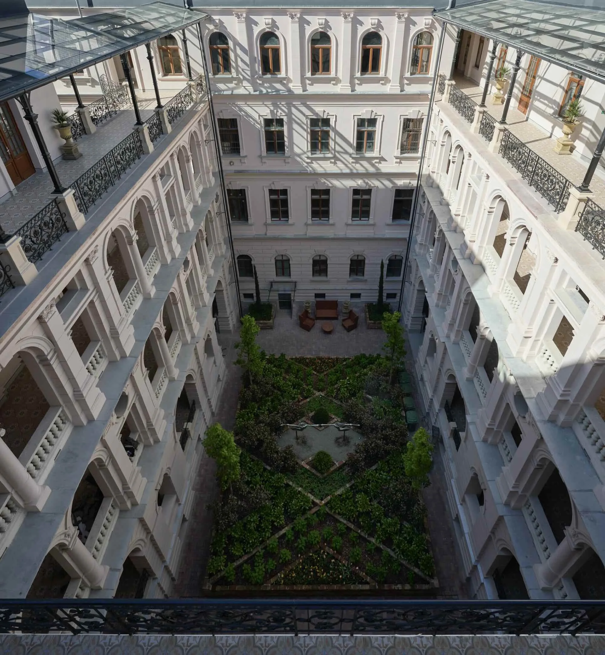 View of a historic-style courtyard garden surrounded by a multi-story white building with arched windows and decorative railings, seen from a balcony.