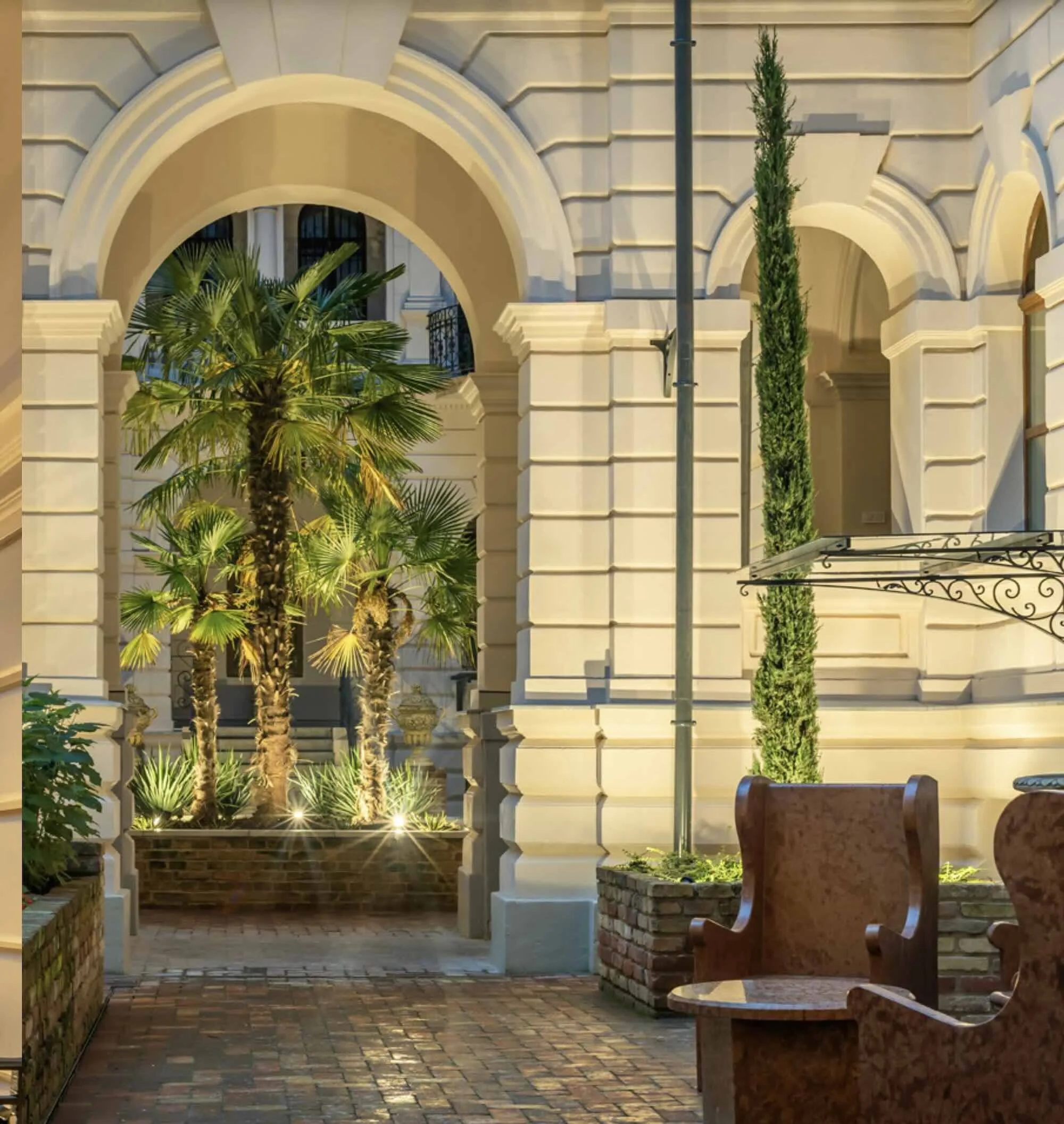 Exterior view of a historic building with arched entryway, illuminated palm trees, and a brick courtyard with wooden chairs.