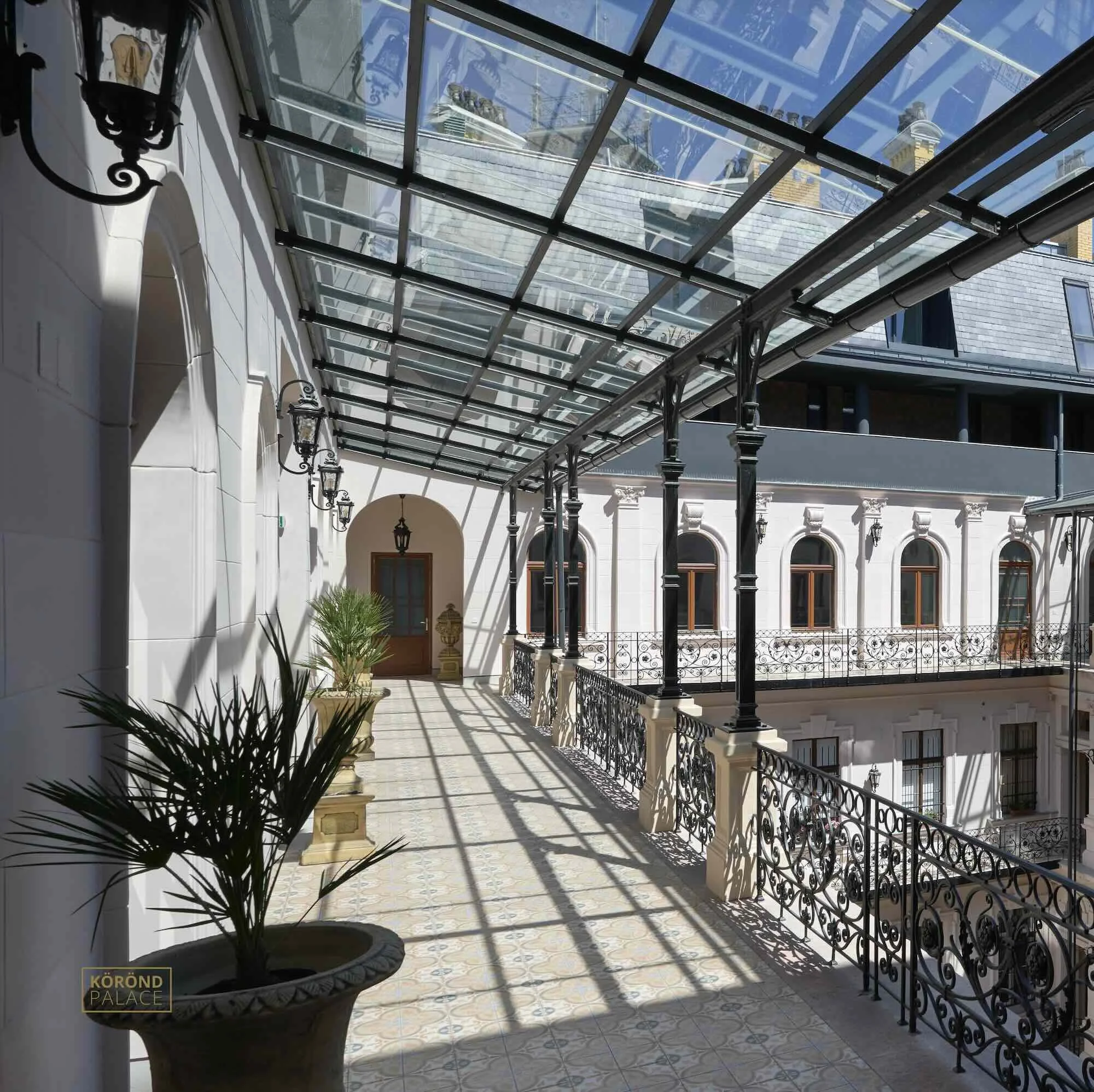 A balcony with ornate black wrought iron railings, potted plants, and a glass roof casting shadows on the floor. The balcony is attached to a historic white building with arched windows and decorative architectural details.