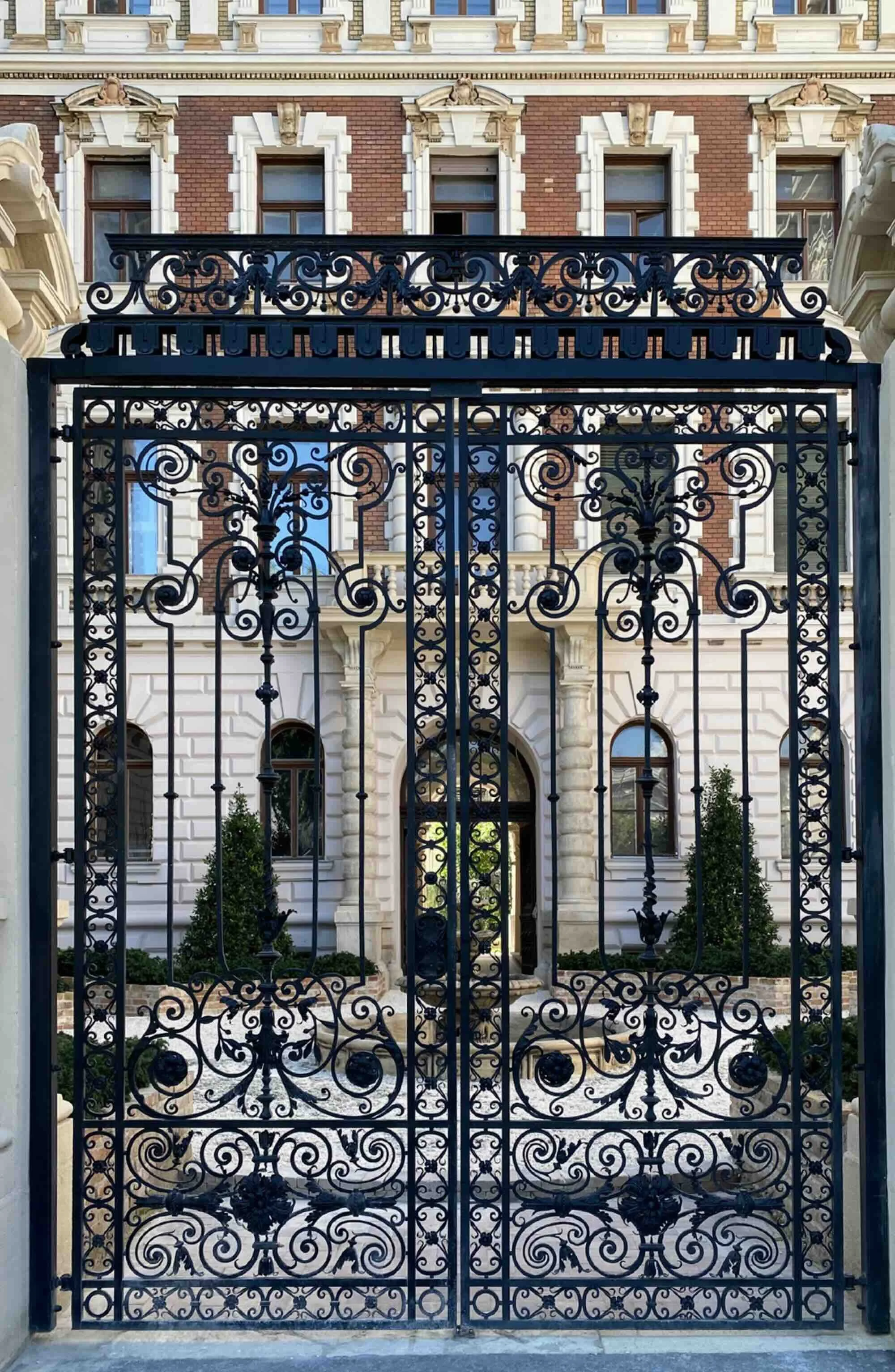 An ornate black iron gate leading to a historic brick and stone mansion with arched windows and greenery in the front yard.