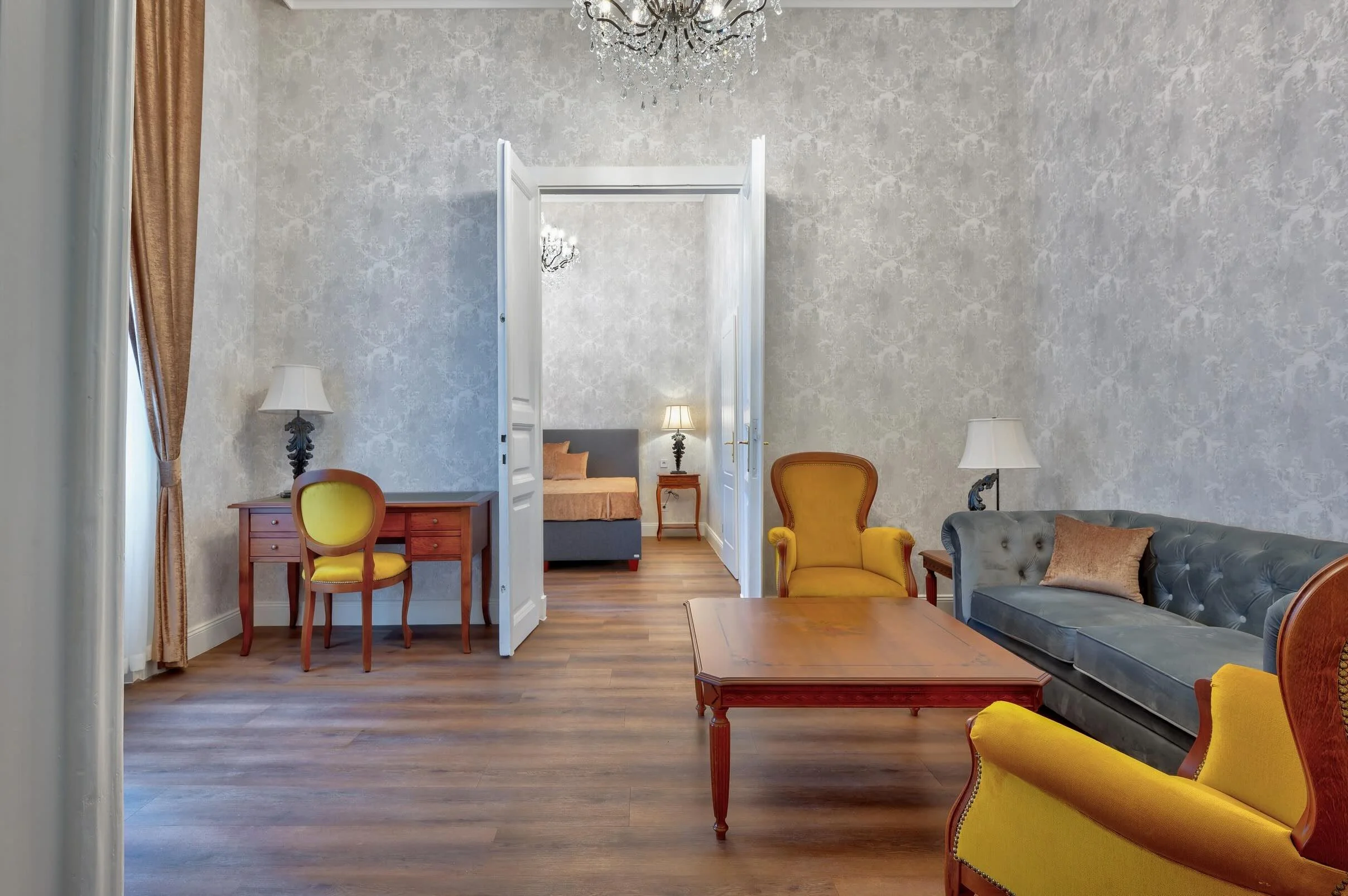 Living room with yellow and gray armchairs, a wooden coffee table, a gray sofa with a decorative pillow, a side table with a lamp, and curtains on the window. The room leads into a bedroom visible through open double doors.