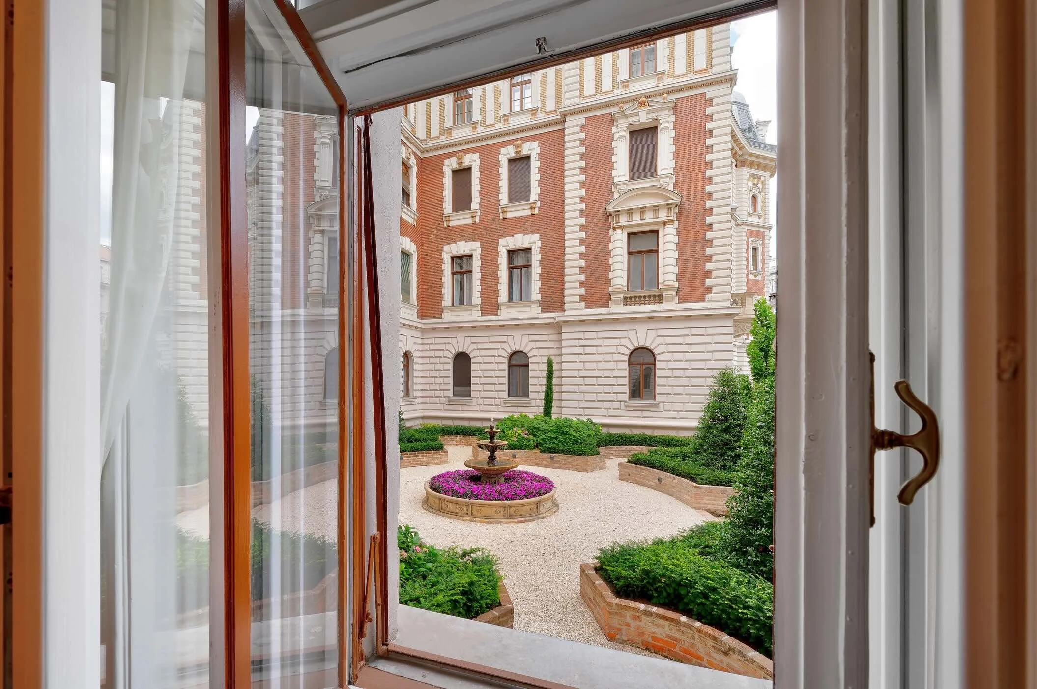 View through a window showing a landscaped courtyard with a fountain, flowers, and greenery, surrounded by tall historic brick and stone buildings.