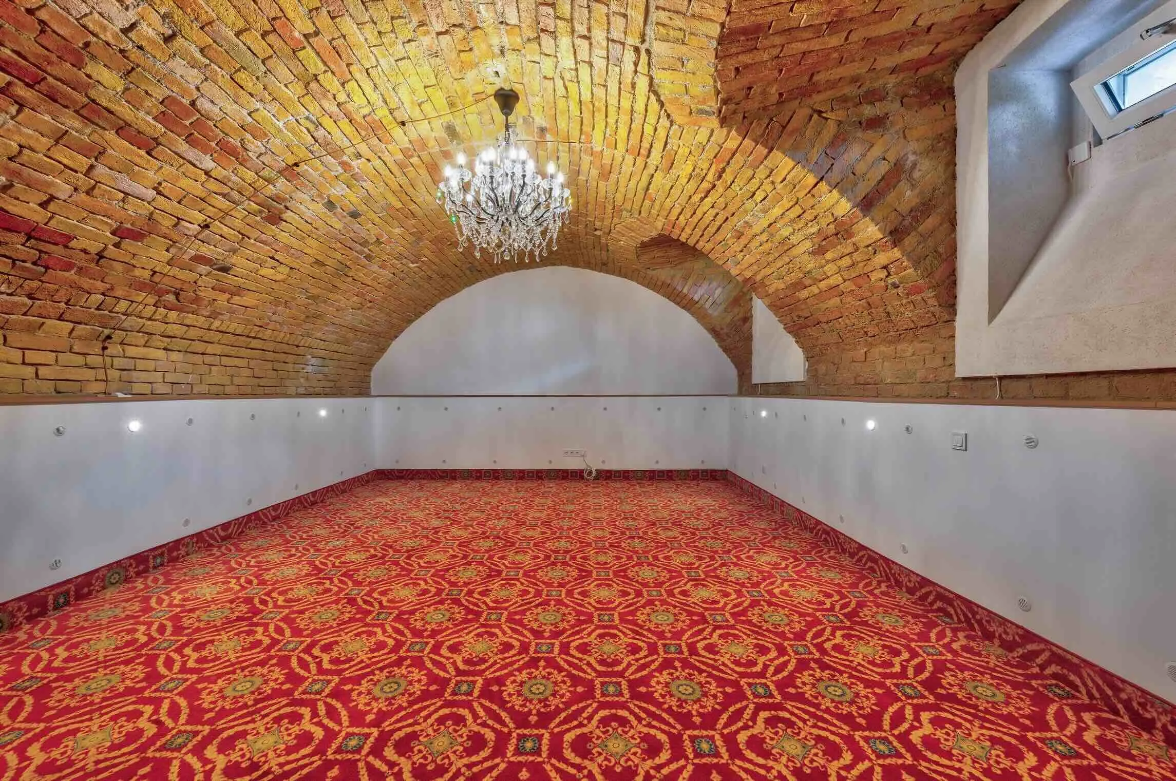 Empty room with arched brick ceiling, chandelier, and a red patterned carpet.