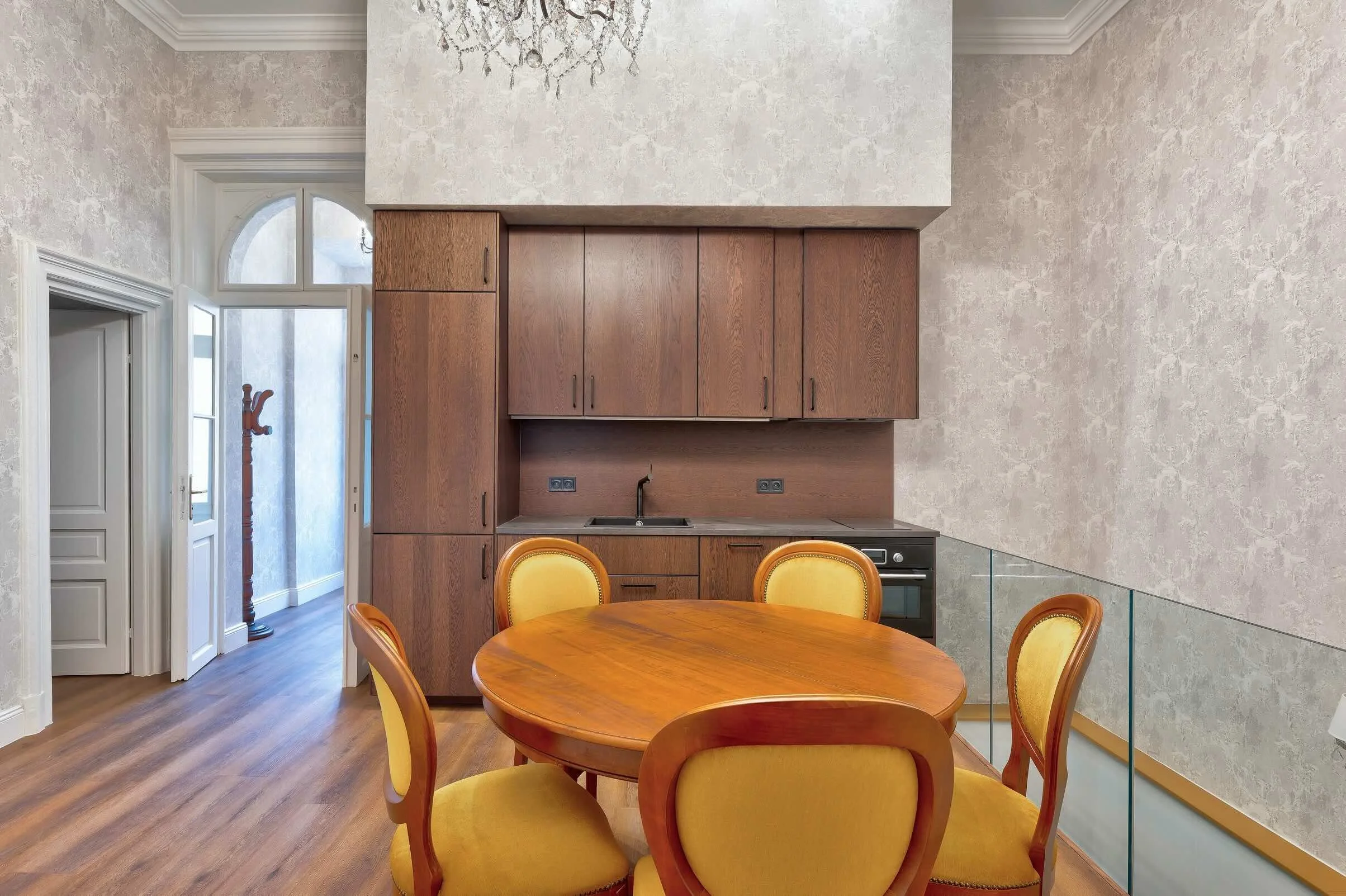 Interior view of a dining area with a wooden round table and six yellow upholstered chairs, a kitchen with wooden cabinets and a built-in oven, and a hallway with a coat stand and arched window.