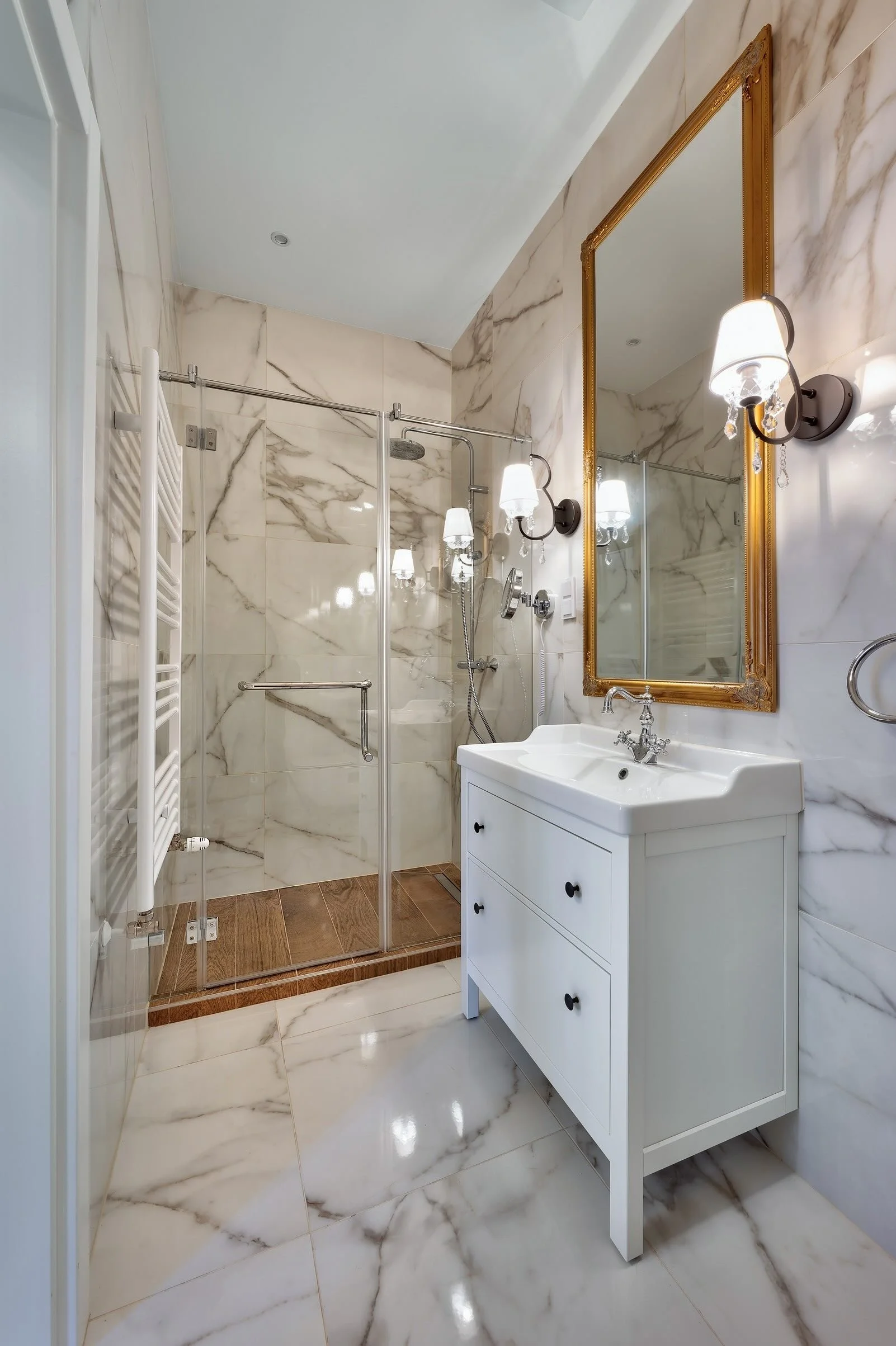 A luxurious bathroom with marble walls and floors, a white vanity with a sink, a large mirror with a gold frame, and a walk-in shower with glass doors and a showerhead.