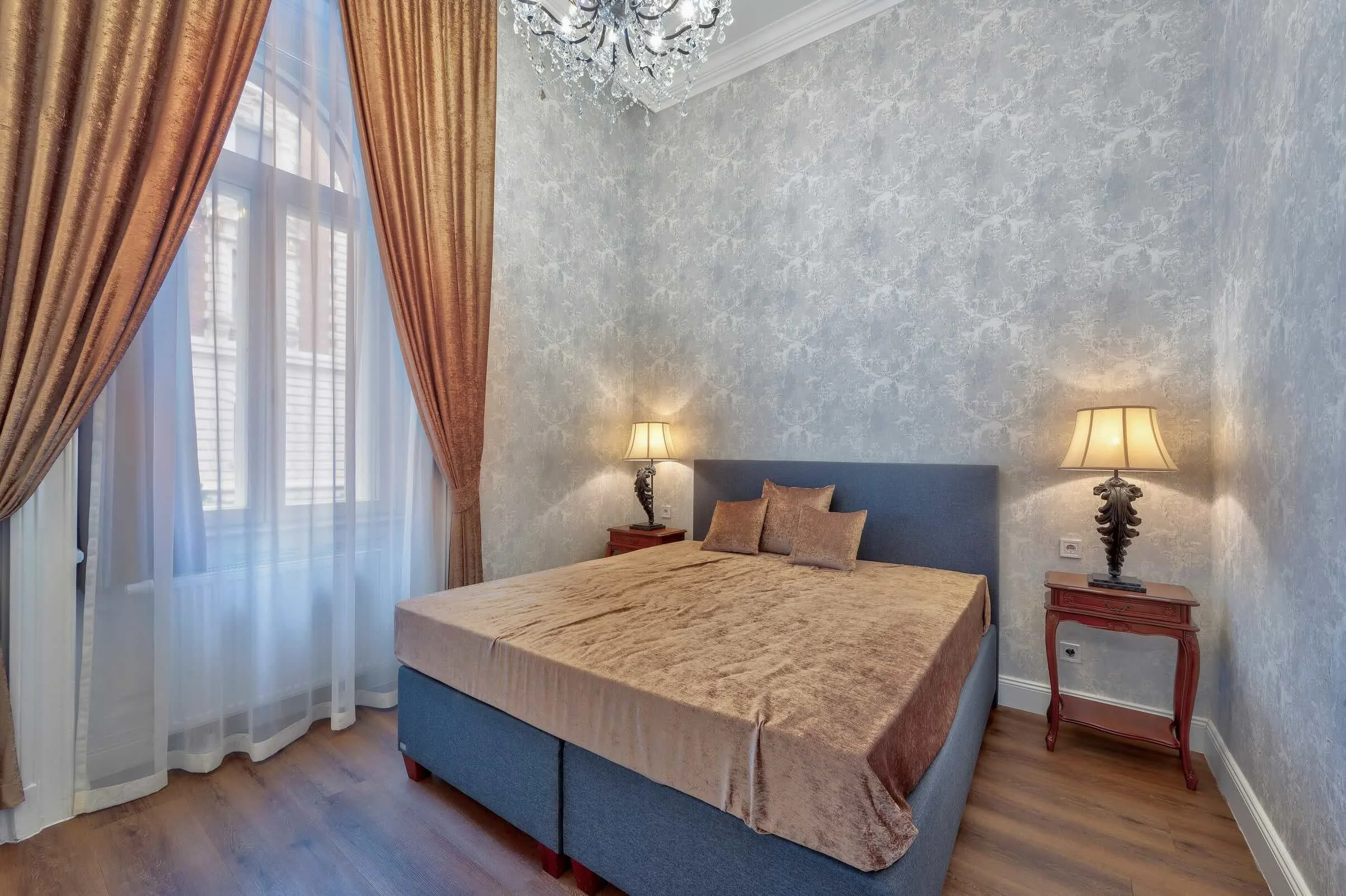 A bedroom with a bed, two nightstands with table lamps, large window with curtains, wooden floor, and textured wallpaper.