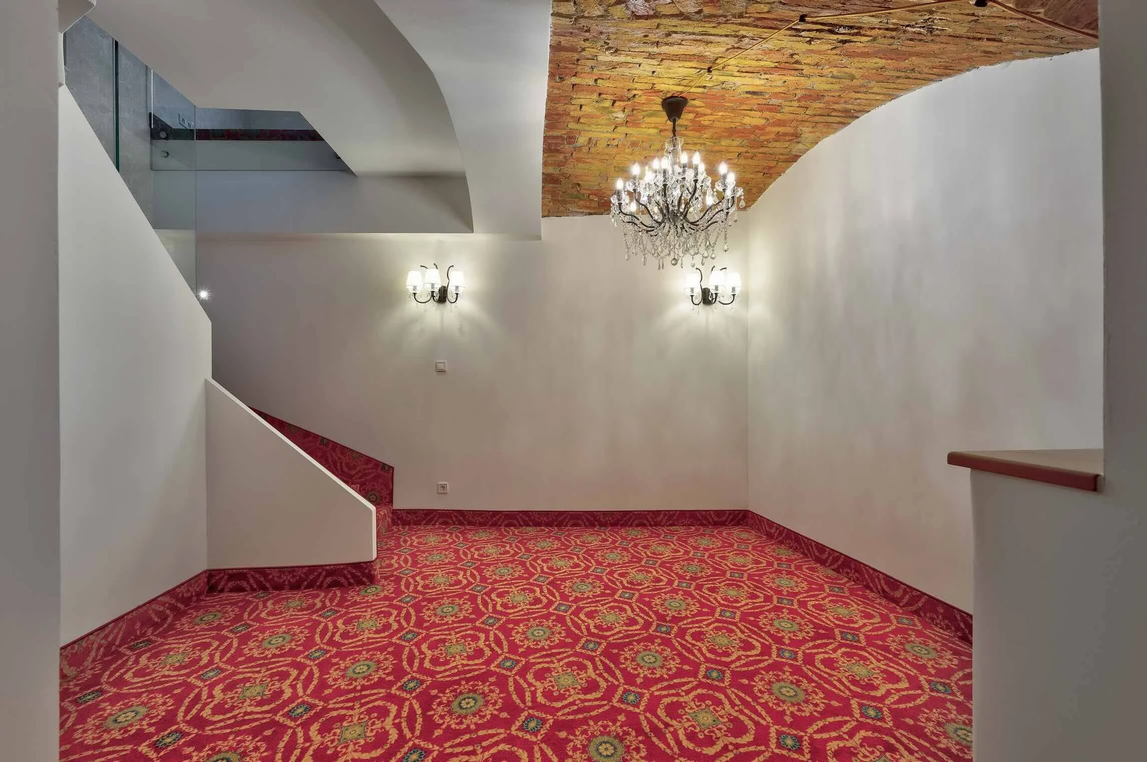 An elegant interior room with white walls, a red patterned carpet, a brick ceiling, and ornate chandeliers and wall sconces lighting.