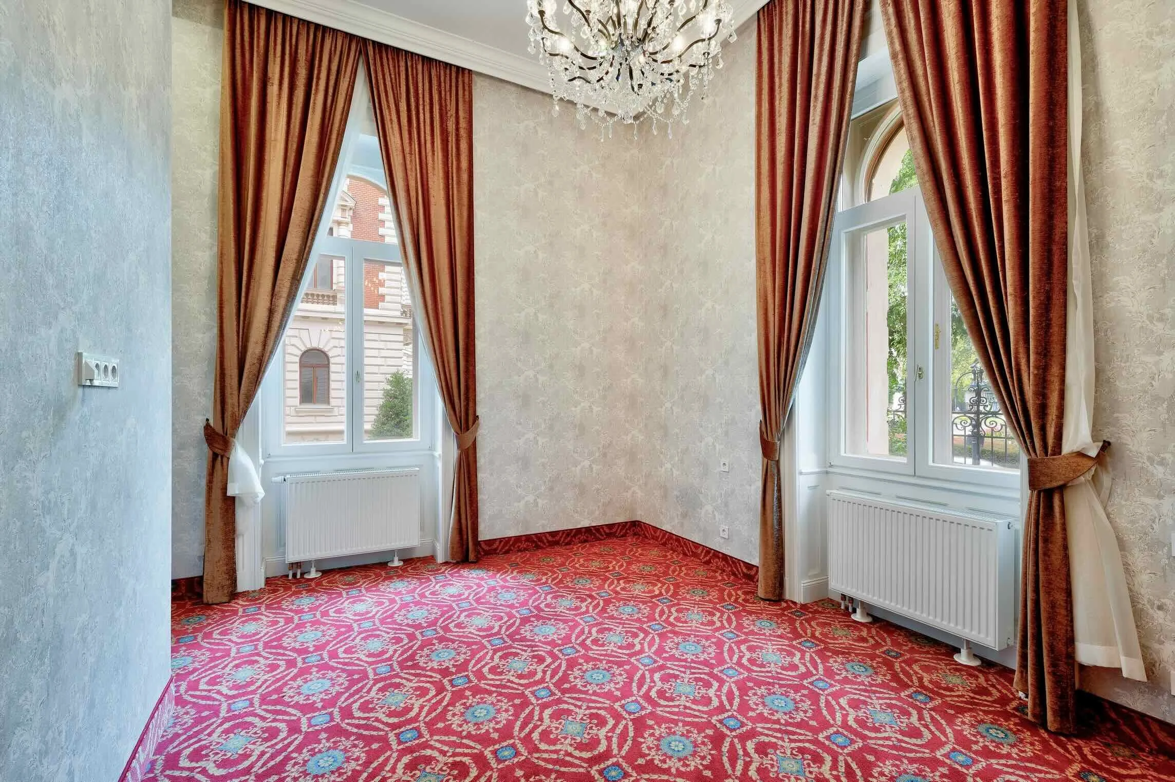 Empty room with ornate red patterned carpet, beige textured wallpaper, and large windows with brown curtains and white trim. A ceiling chandelier hangs from the ceiling.