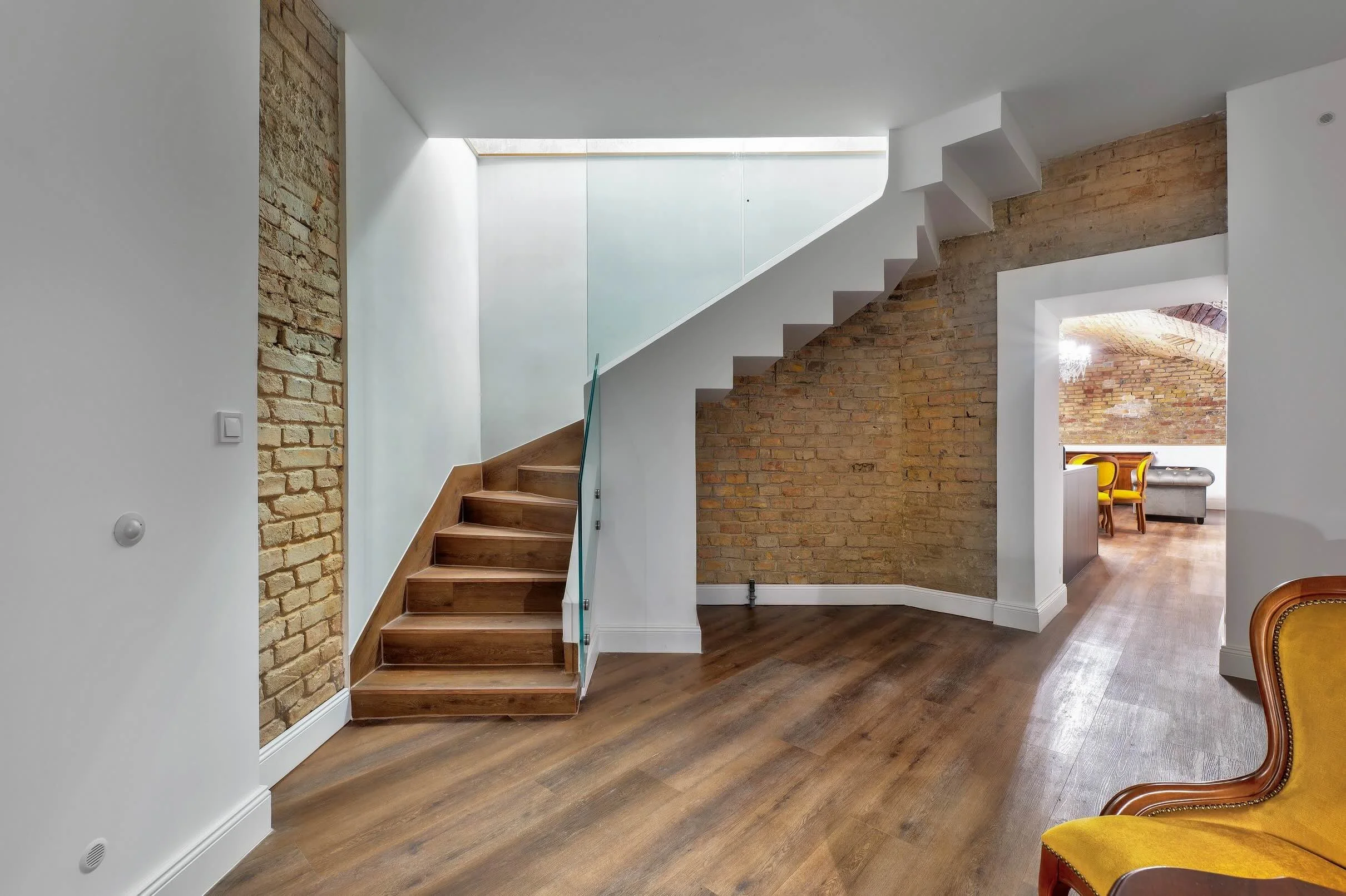 Interior of a modern home with wooden floors, brick walls, a staircase with glass railing, and a view into another room with chairs and a chandelier.