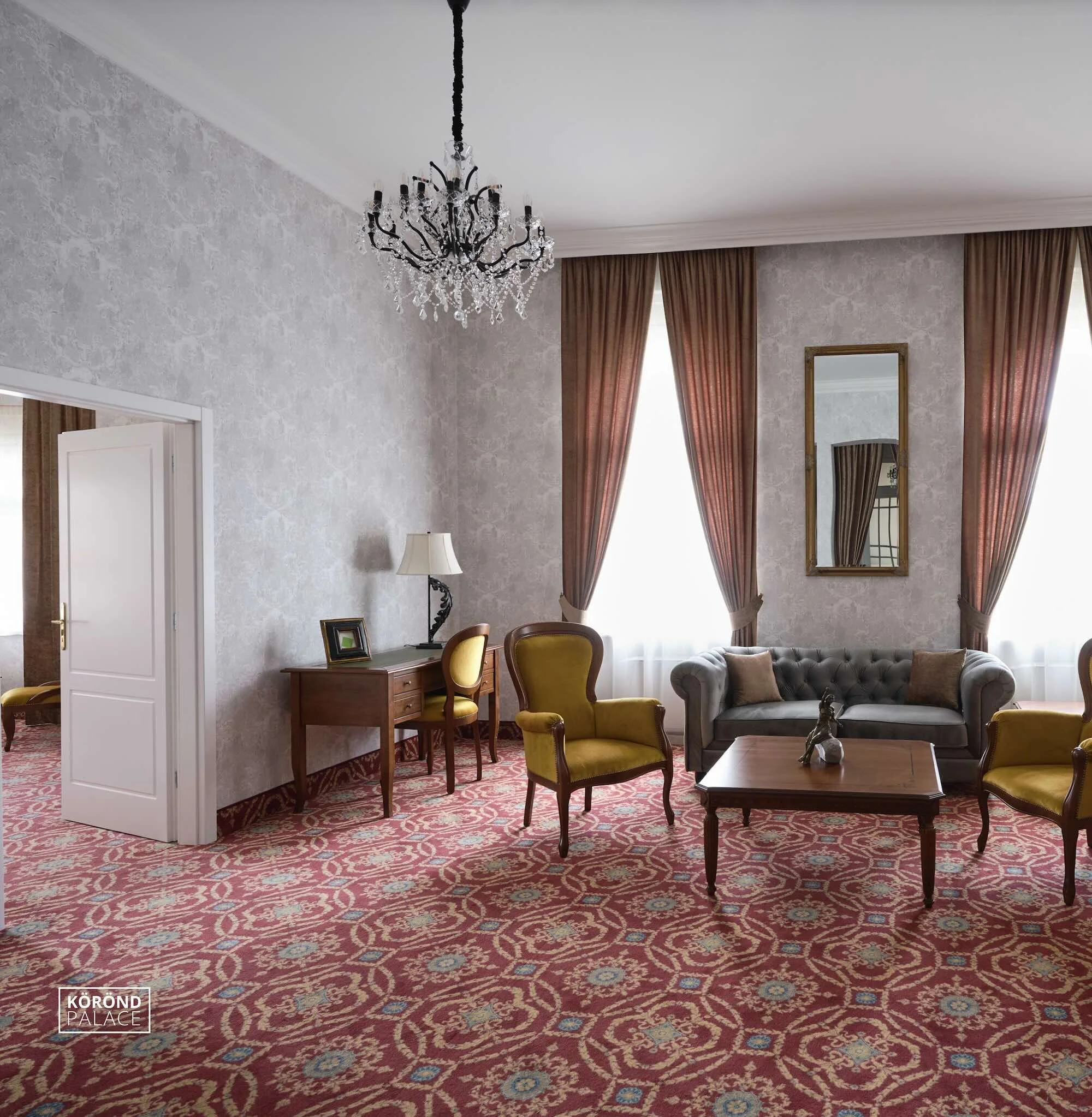 Elegant living room with a chandelier, patterned red carpet, velvet armchairs, grey tufted sofa, wooden coffee table, framed mirror, table lamp, and large windows with draped curtains.