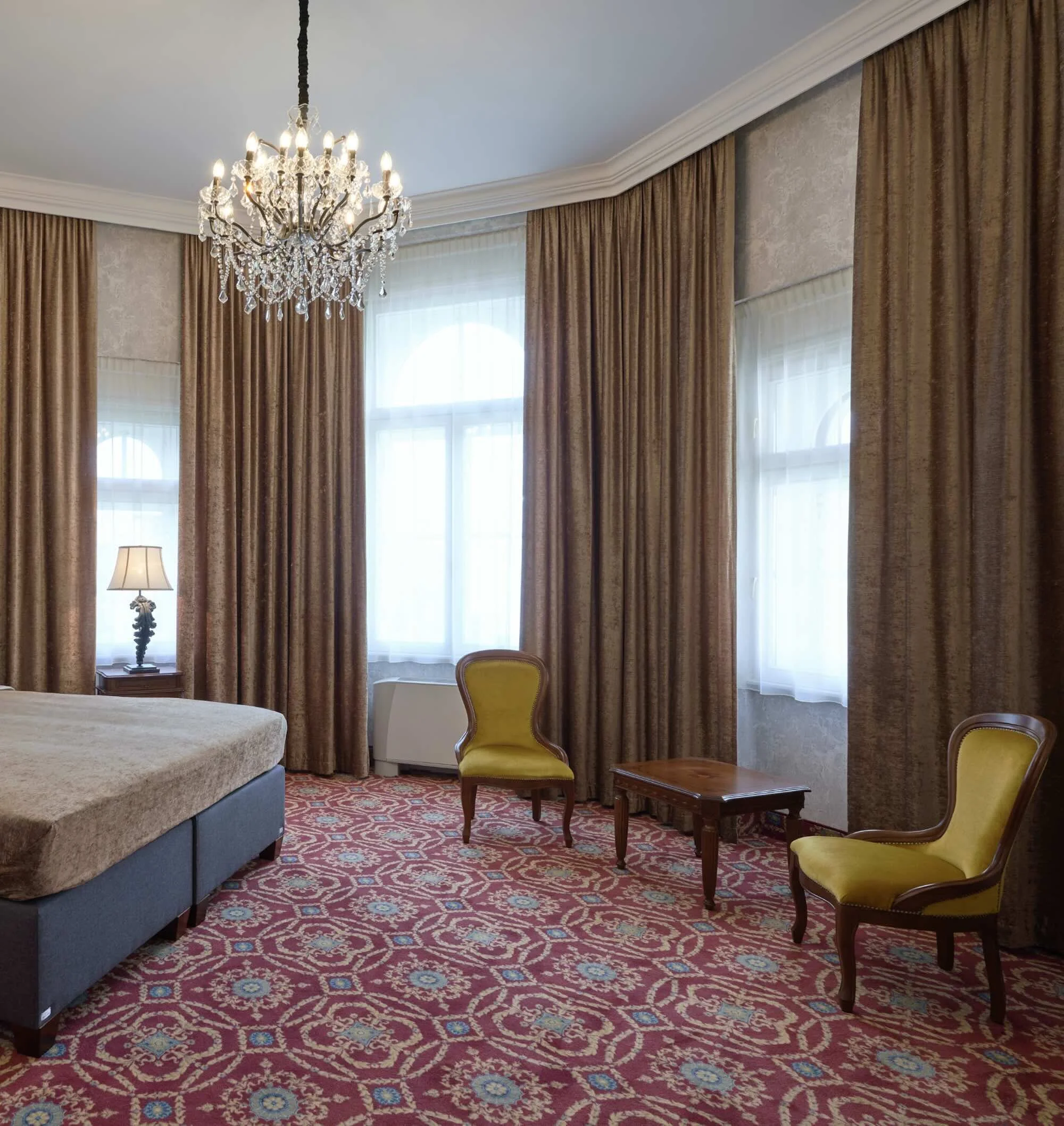Elegant hotel room with large windows, beige curtains, crystal chandelier, yellow armchairs, wooden side table, patterned red carpet, bed partially visible, lamps, and traditional decor.