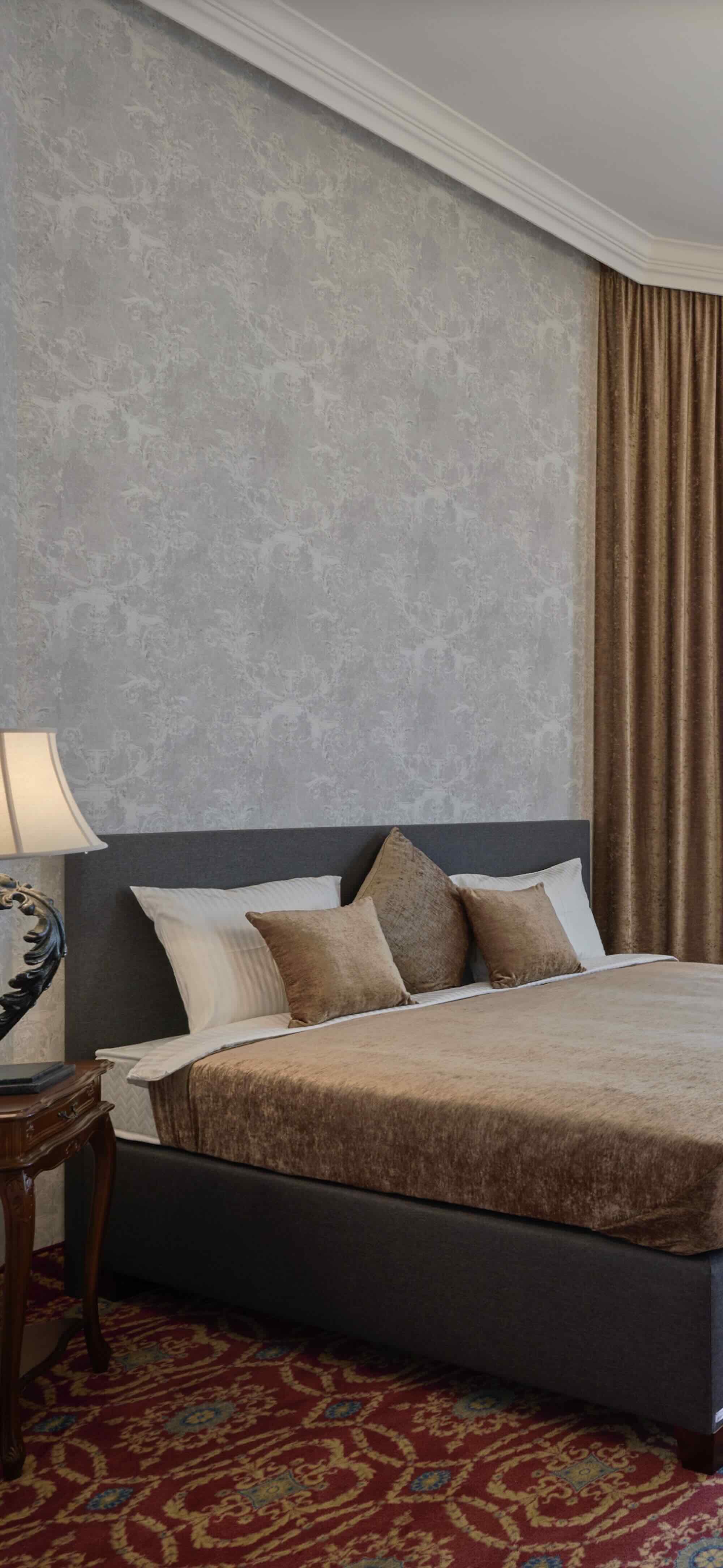 A hotel room with a bed, pillows, a bedside table with a lamp, beige and gold curtains, a patterned carpet, and an ornate ceiling molding.