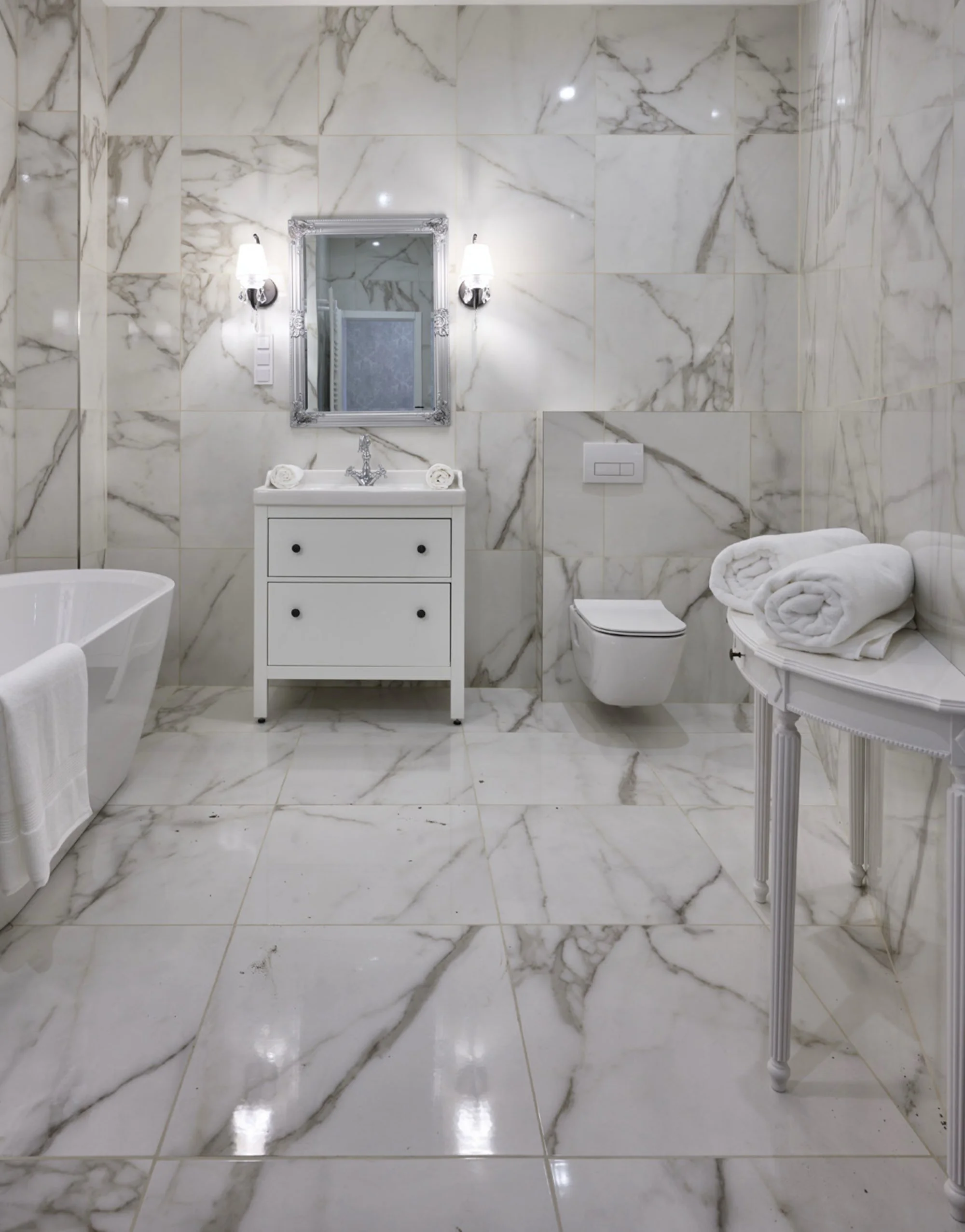 A luxurious bathroom with white marble walls and floor, featuring a bathtub, a white vanity with a mirror, a wall-mounted toilet, and a small table holding rolled white towels.