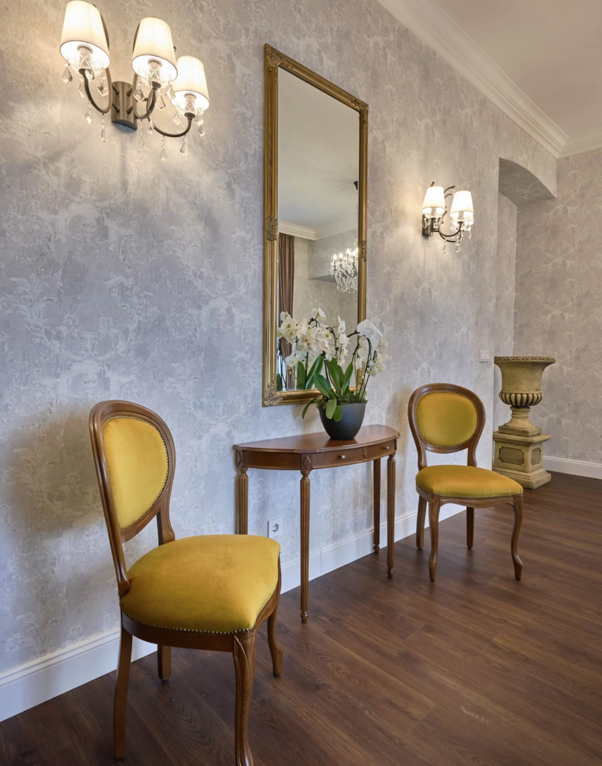 Elegant hallway with two yellow upholstered chairs, a narrow wooden table, a large mirror, floral arrangement, wall-mounted lamps, and a decorative planter.