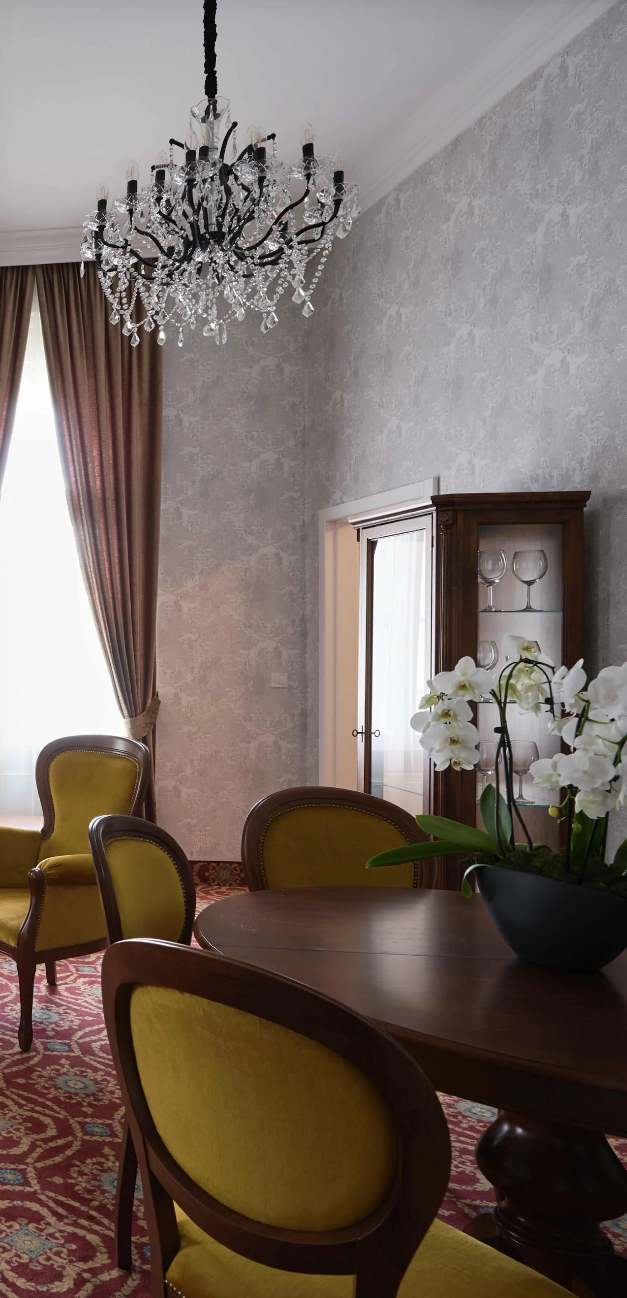 Interior of a dining room with a chandelier, yellow upholstered chairs, a wooden table, a floral arrangement, and a glass display cabinet with wine glasses, featuring gray textured walls, heavy brown curtains, and a patterned rug.